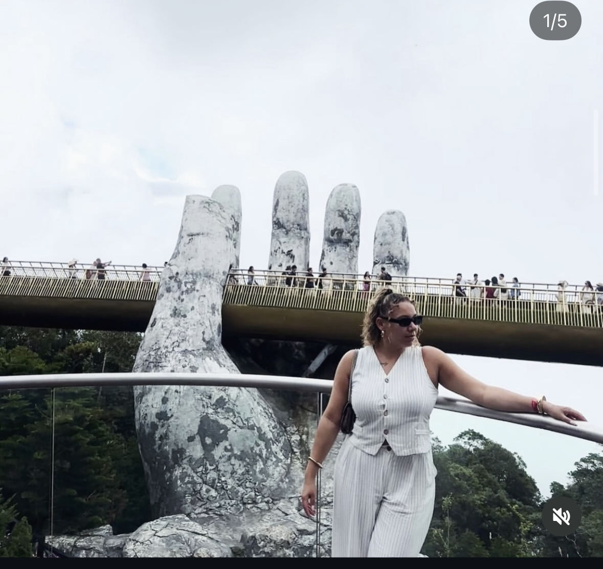 A woman wearing a white striped sleeveless outfit and sunglasses touching a railing in front of the Golden Bridge in Vietnam, with giant stone hands holding up the bridge and visitors walking along it.