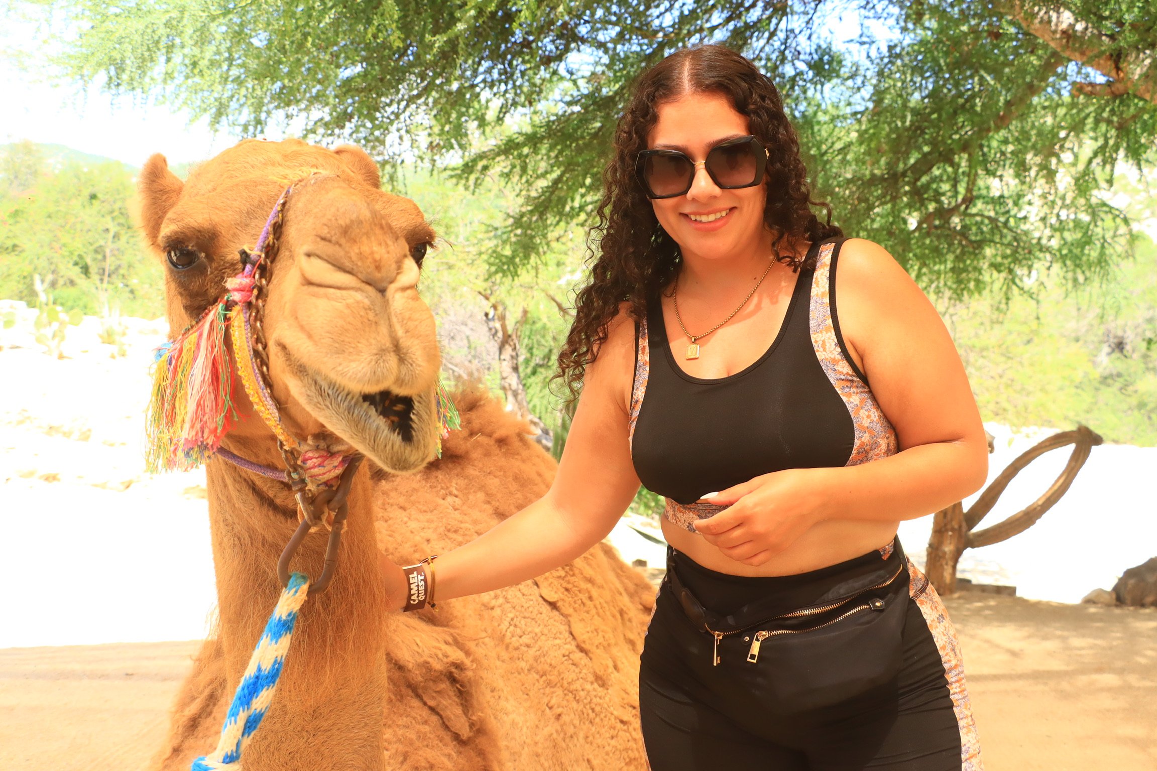 Woman smiling and petting a camel outdoors on a sunny day, with green trees in the background.