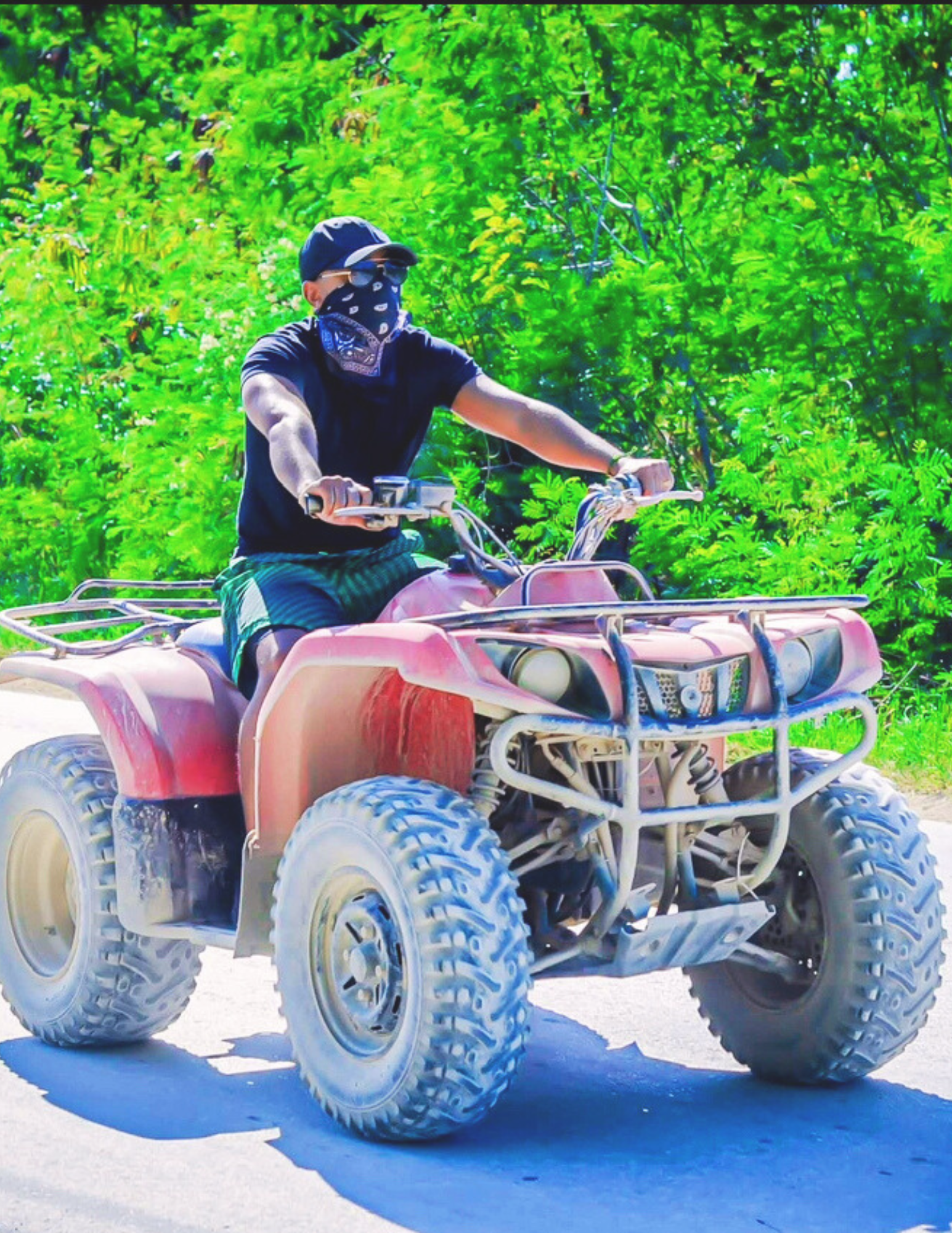Person wearing black shirt, sunglasses, and bandana riding a red all-terrain vehicle on a sunny day with green trees in the background.