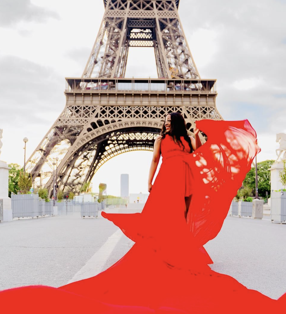 A woman in a flowing red dress standing in front of the Eiffel Tower in Paris, smiling and holding part of her dress as it billows in the wind.