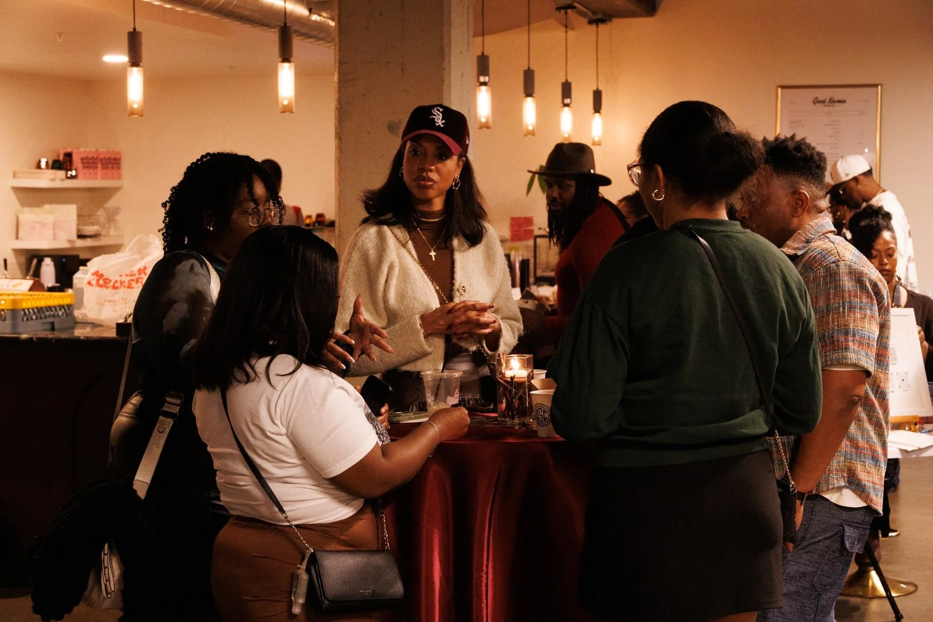 A diverse group of people gathered around a high table inside a warmly lit cafe or restaurant, engaged in conversation.