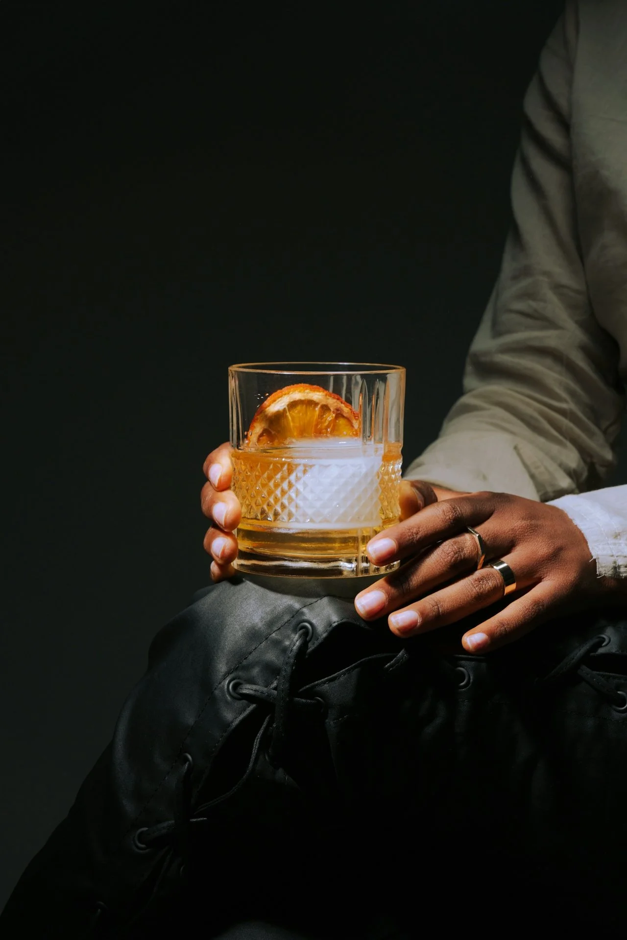 Person holding a glass of whiskey with an orange slice garnish, wearing rings, sitting on a black surface against a dark background.