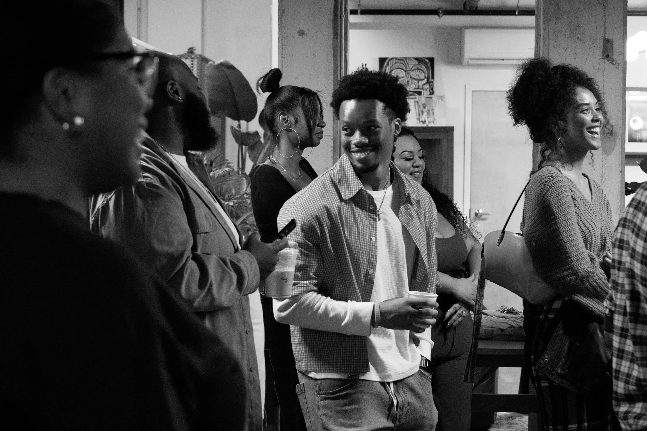 Group of diverse people smiling and socializing indoors, some holding drinks, in a modern casual setting.