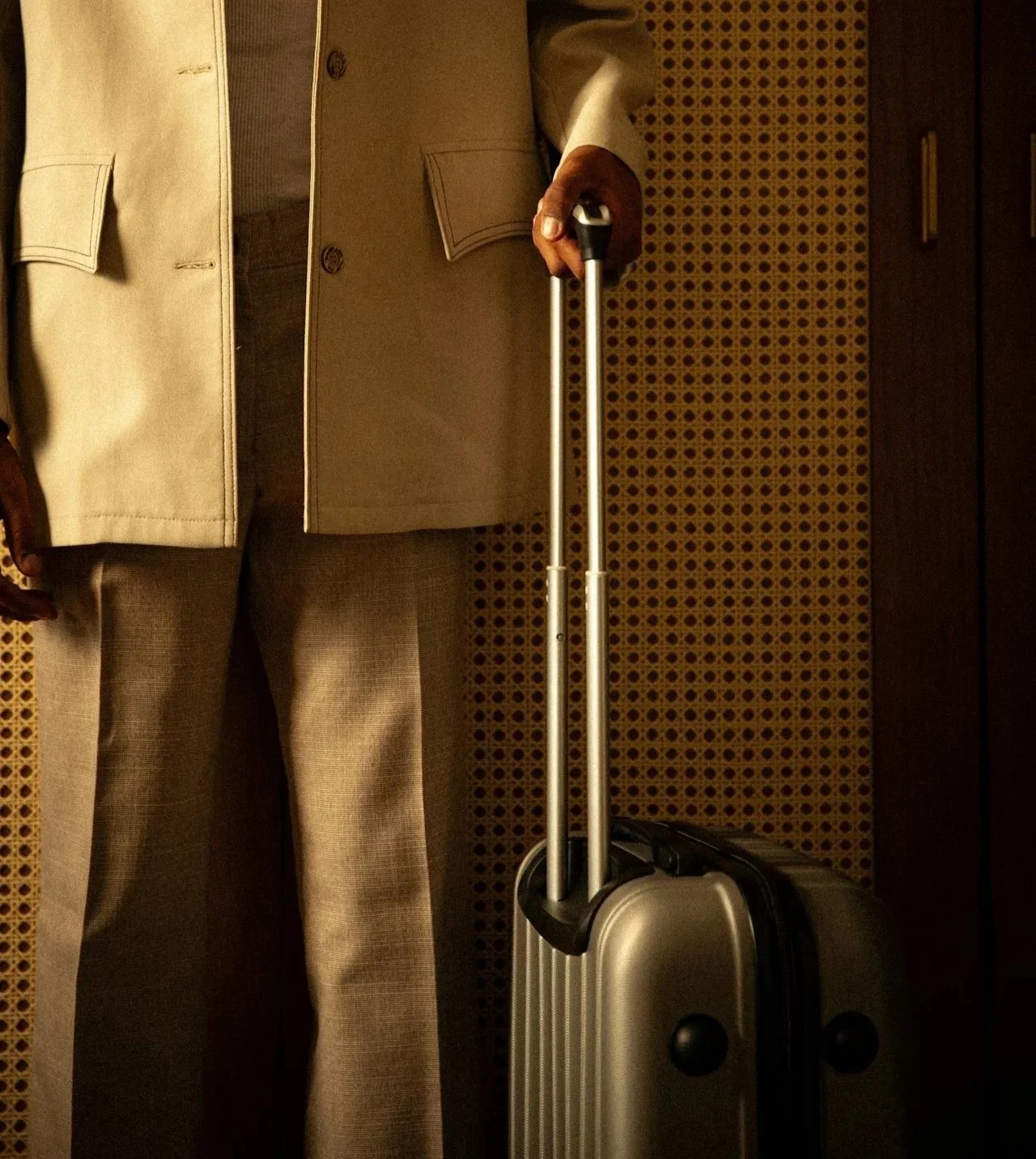 Person standing in a beige suit holding a silver rolling suitcase with an extended handle.