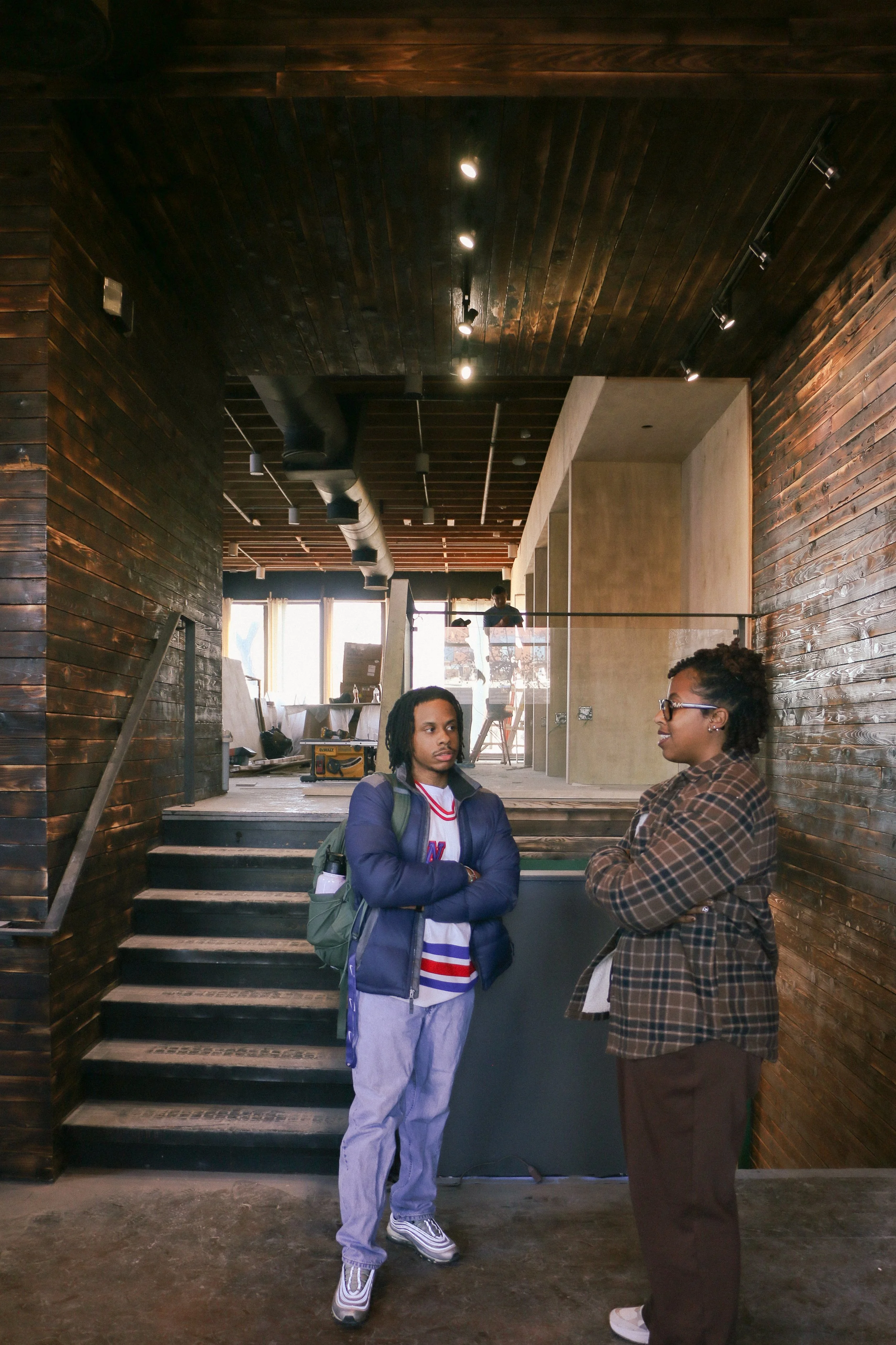 Two people having a conversation inside a modern building with wooden walls and ceiling. One person is wearing a striped shirt and glasses, the other is wearing a jacket and backpack.