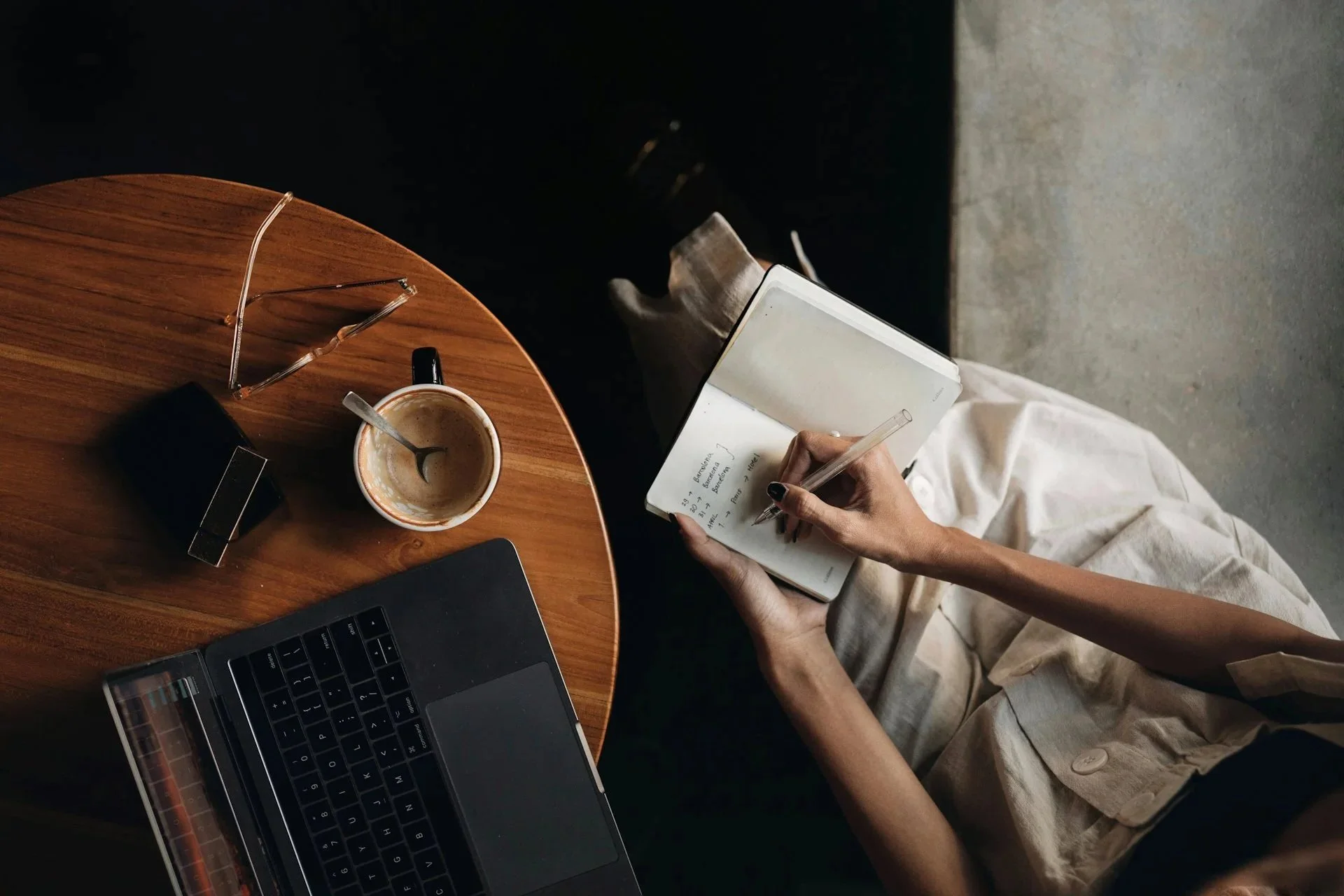 A person sitting at a wooden table with a laptop, a cup of coffee with a spoon inside, a pair of glasses, and a smartphone. The person is writing in a notebook with a pen.