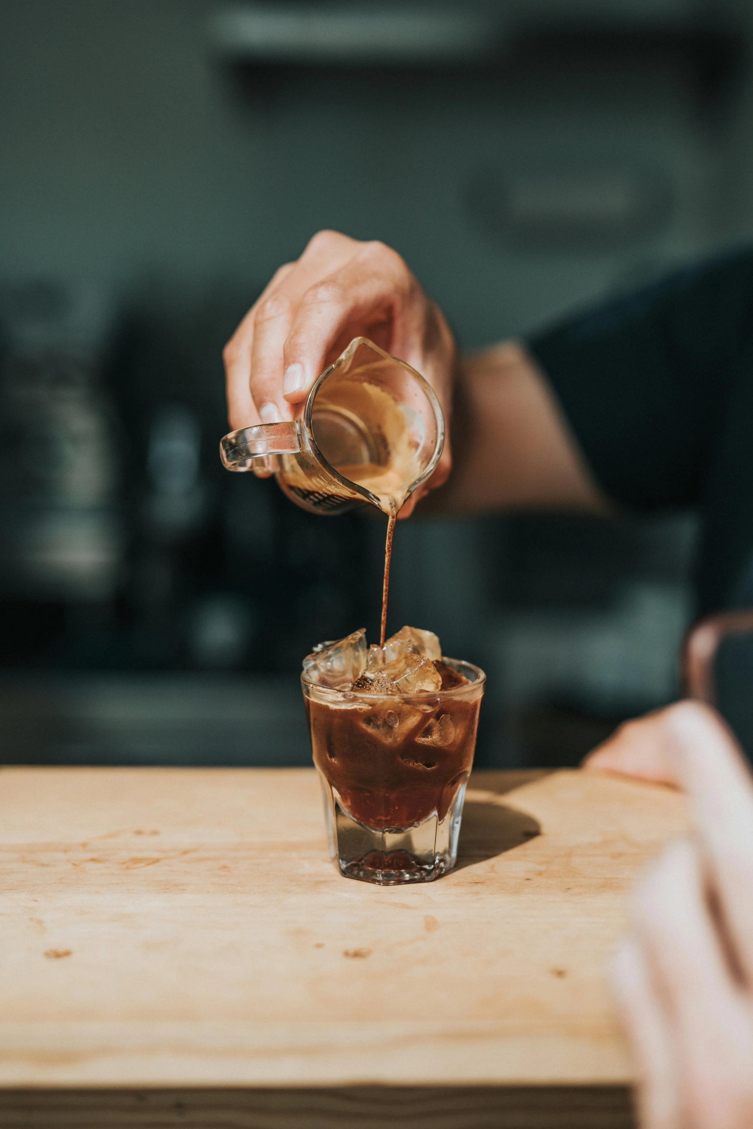 Person pouring coffee into a glass filled with ice on a wooden table.