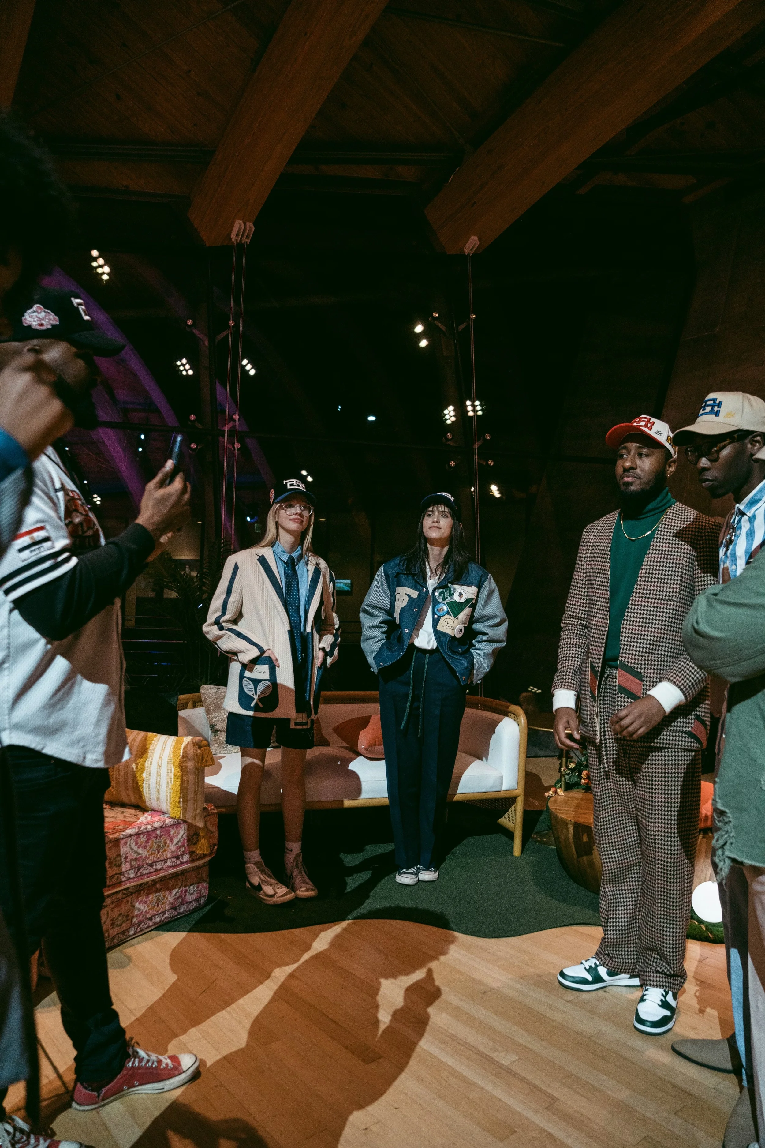 A group of five people standing and talking in a room with wood ceiling beams, in front of a sofa with pillows and a small round table. The scene appears to be indoors under low lighting.