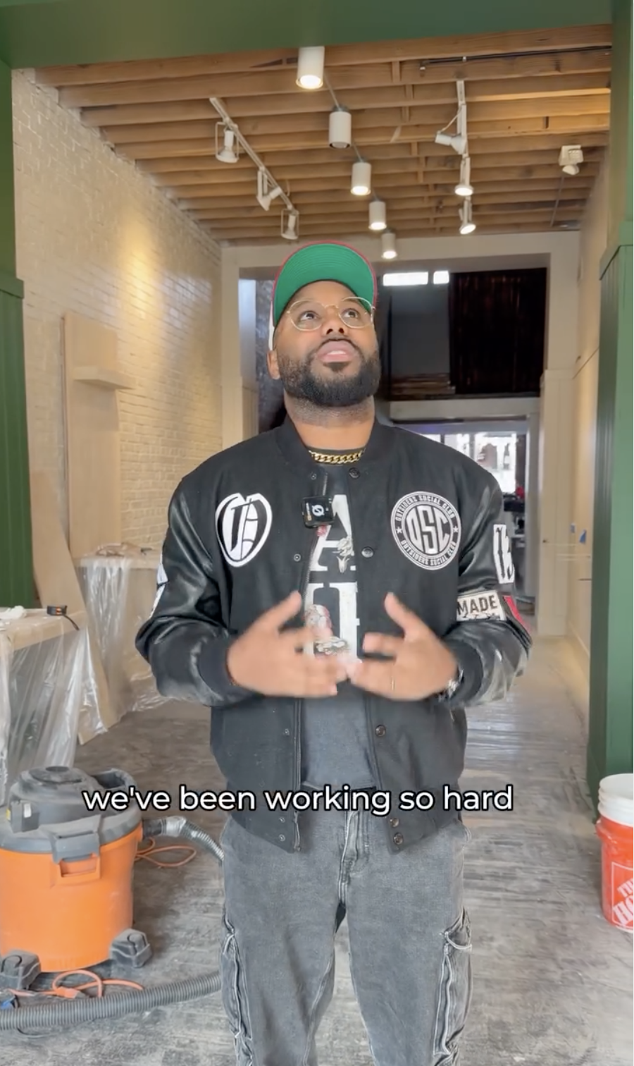 Man with a beard wearing glasses, a black jacket, a gray shirt, a red and green cap, and a gold chain speaking inside a room under construction, with a vacuum cleaner and unfinished walls around him.