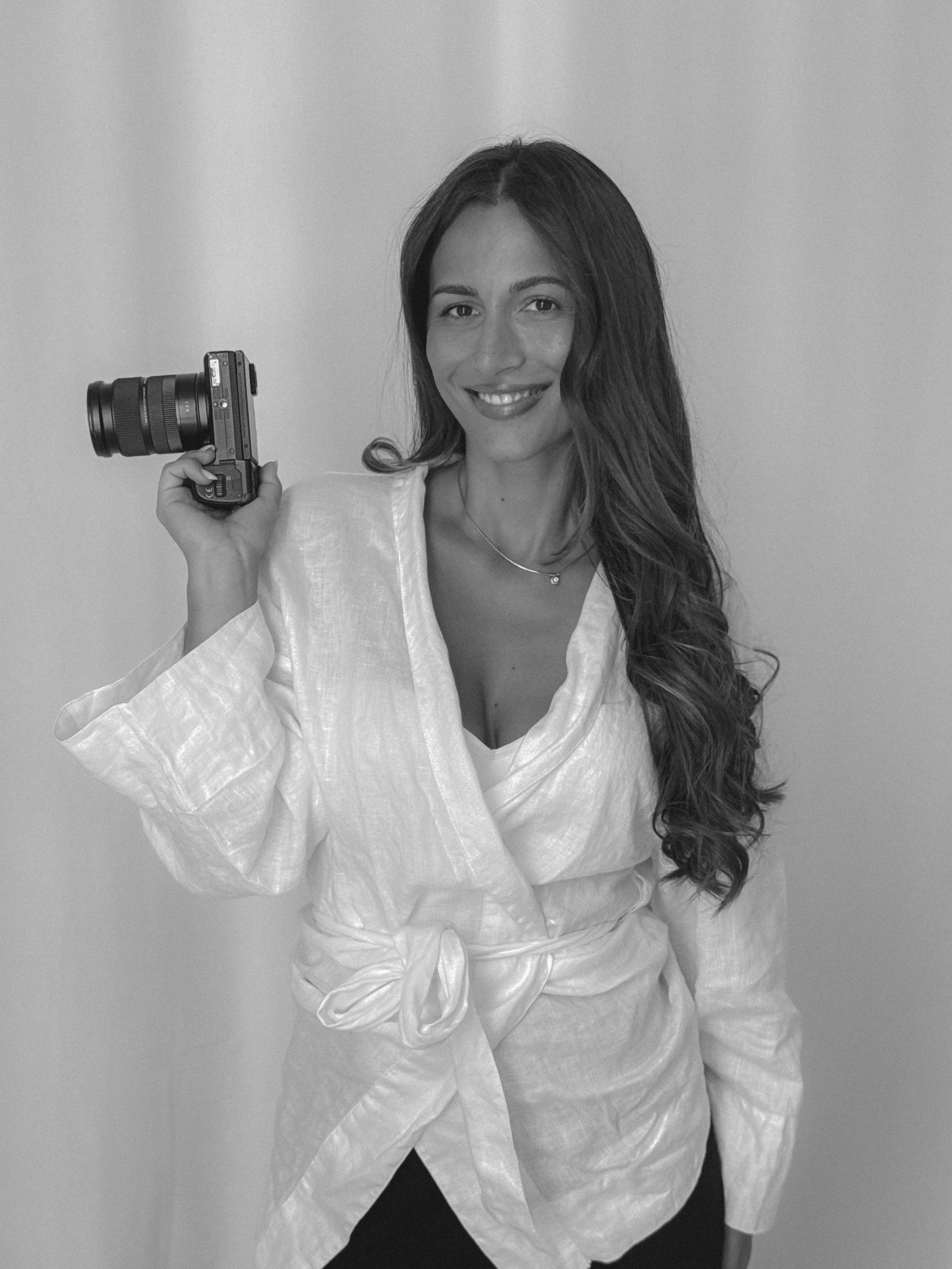 Mujer sonriendo con cabello largo y oscuro, vestida con ropa blanca y sosteniendo una cámara en su mano derecha.