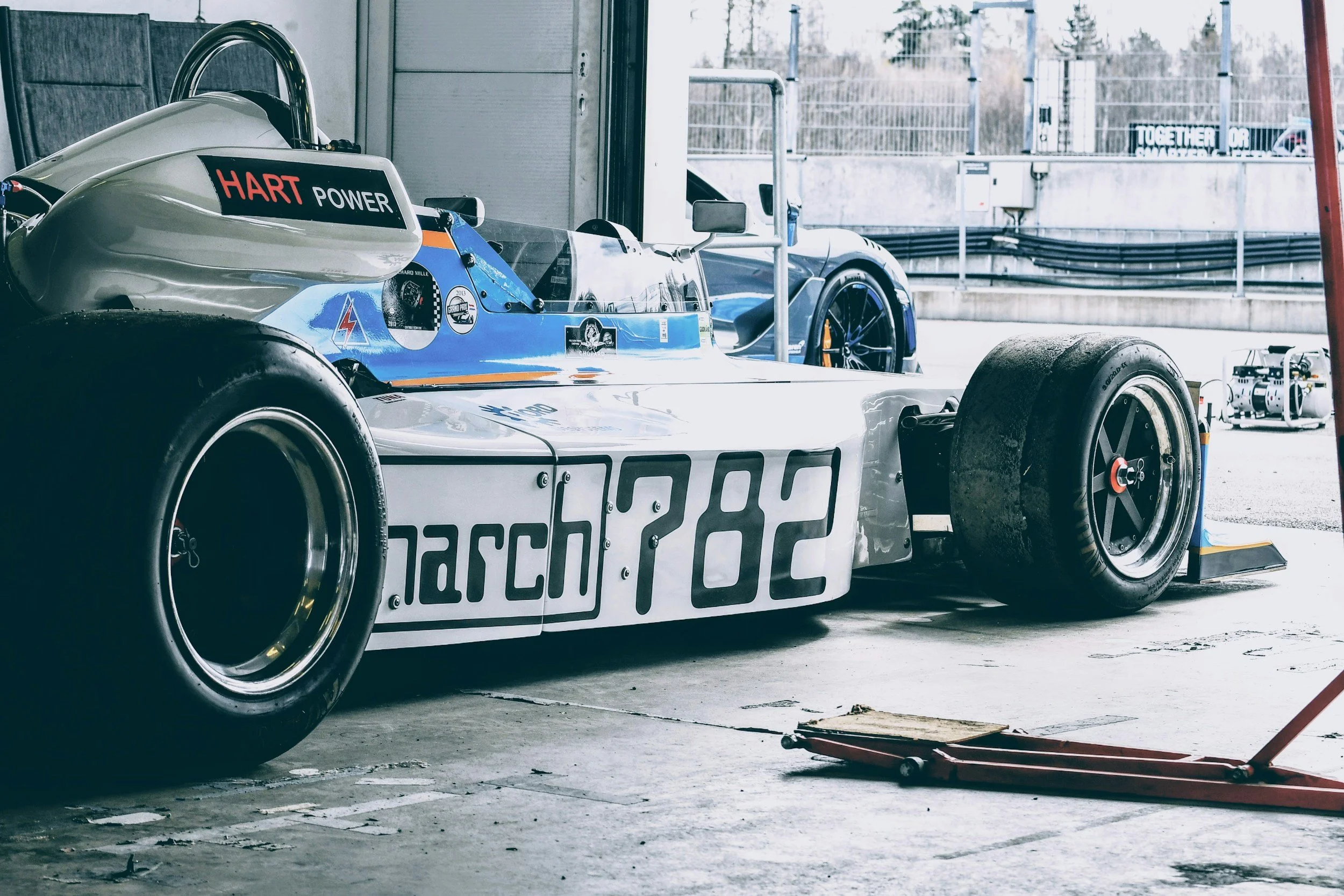 A race car in a garage with a white and blue body, large tires, and sponsorship decals, including 'March 282', 'Hart Power', and others, with another cars visible outside the garage.