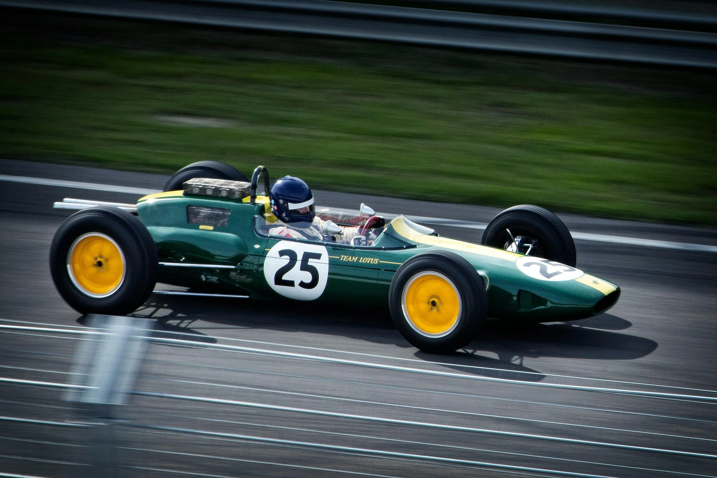 Green vintage racing car with the number 25 on white circles, speeding on a racetrack, driven by a driver wearing a blue helmet and white racing suit, with yellow wheels and chrome accents.