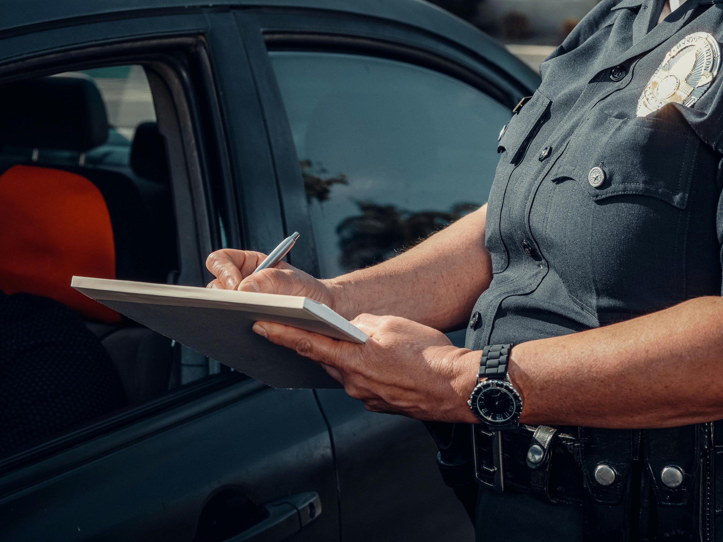 Close-up of a police officer writing a report on a notepad next to a parked police car.