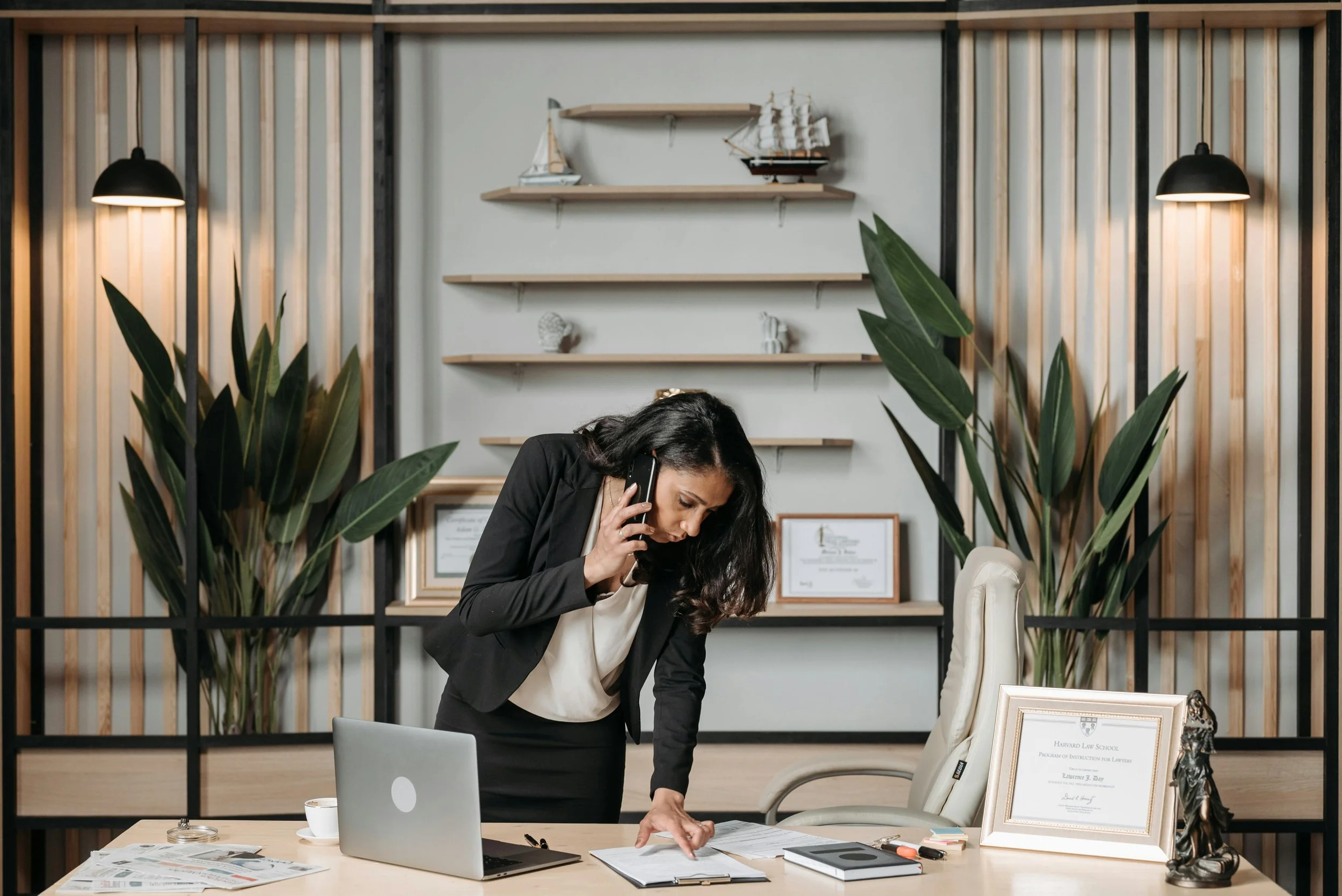 Businesswoman in a modern office, talking on the phone, leaning over a desk with documents, a laptop, and framed certificates, surrounded by large green plants.