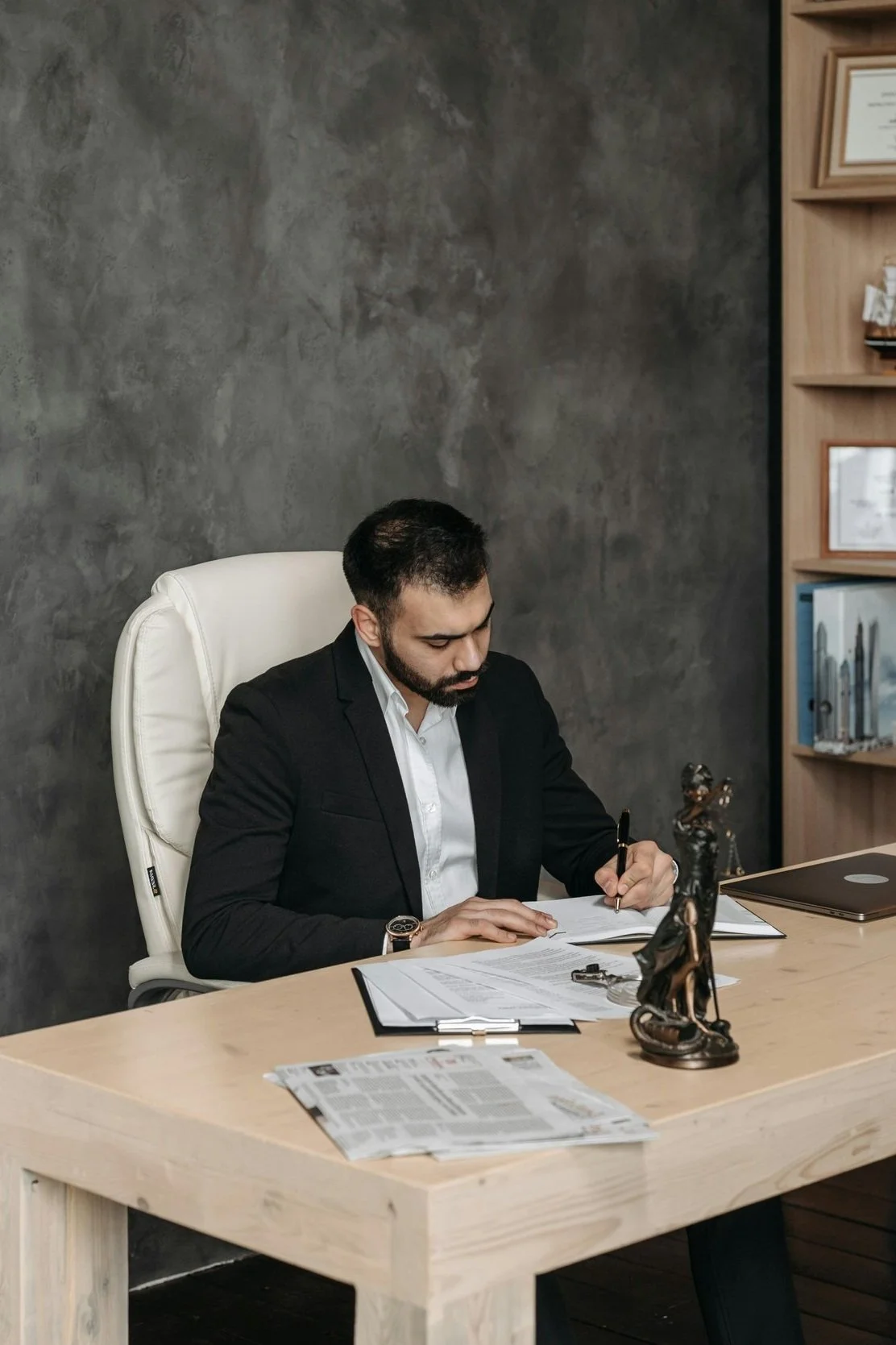 A man in a black suit sitting at a wooden desk, writing on a document with a pen, surrounded by legal papers and decorative items, in an office with a textured gray wall and a bookshelf.