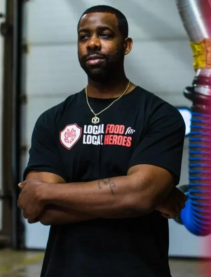 A man with a beard and short hair wearing a black T-shirt with a logo and printed text, crossing his arms, standing indoors.