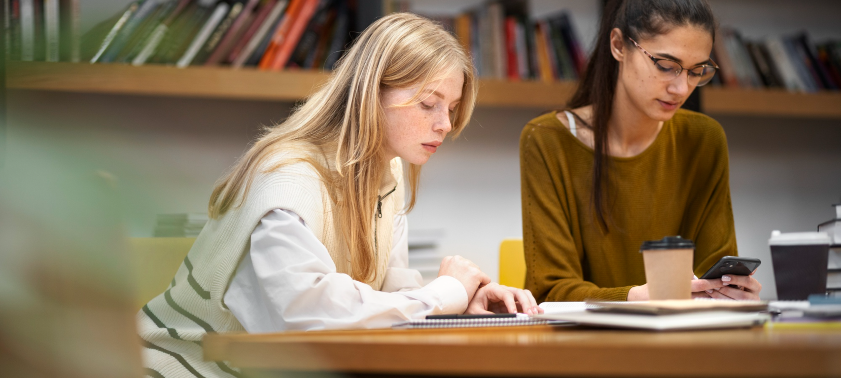 Two women studying at a table in a library or bookstore, with bookshelves in the background. One woman has blonde hair, wearing a white and cream vest, and is reading or writing. The other woman has dark hair, glasses, a brown top, and is looking at her phone.