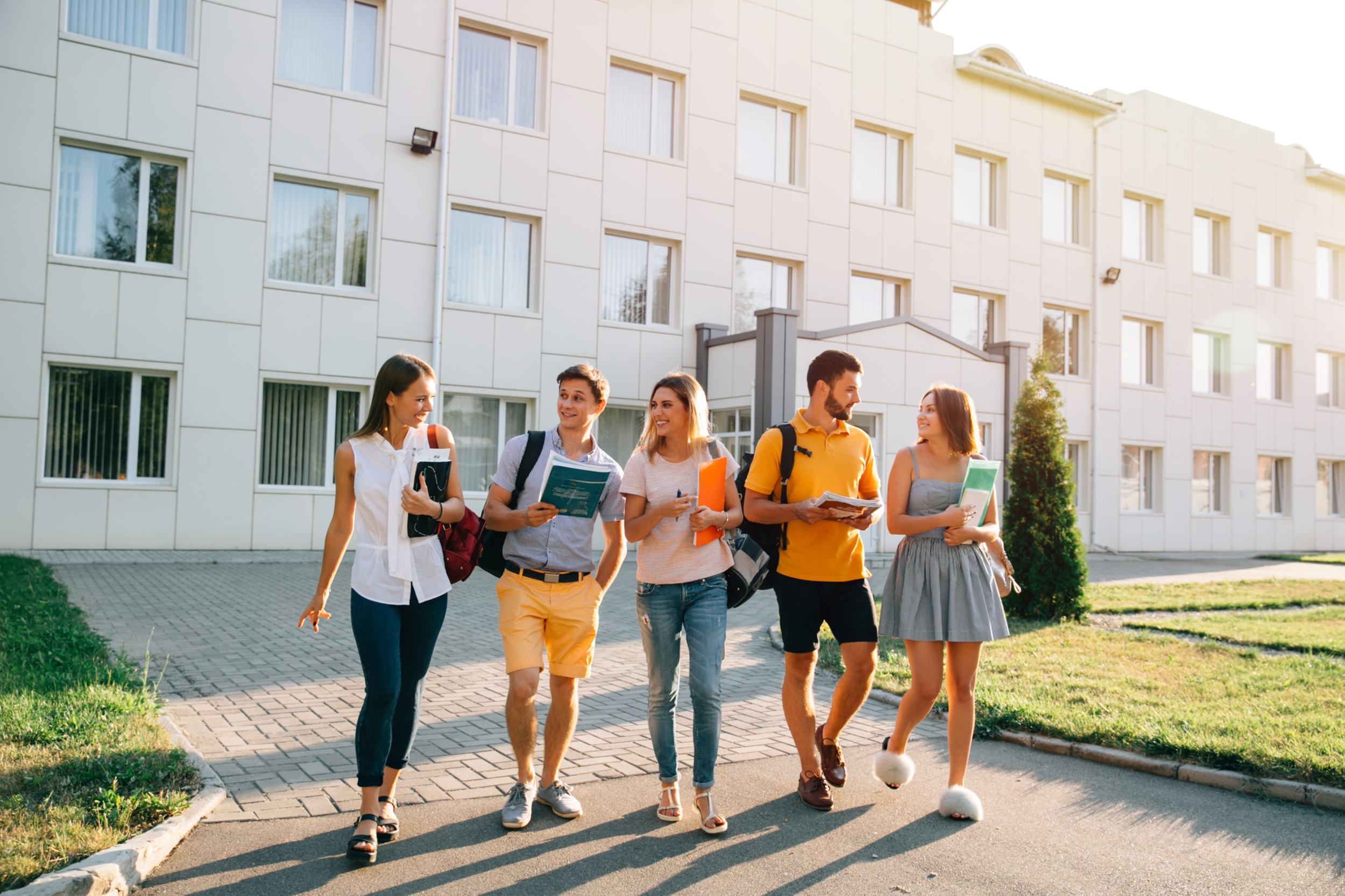 A group of five young adults walking outside in front of a modern multi-story residential building, carrying books, notebooks, and backpacks, engaged in conversation and smiling, with sunlight illuminating the scene.
