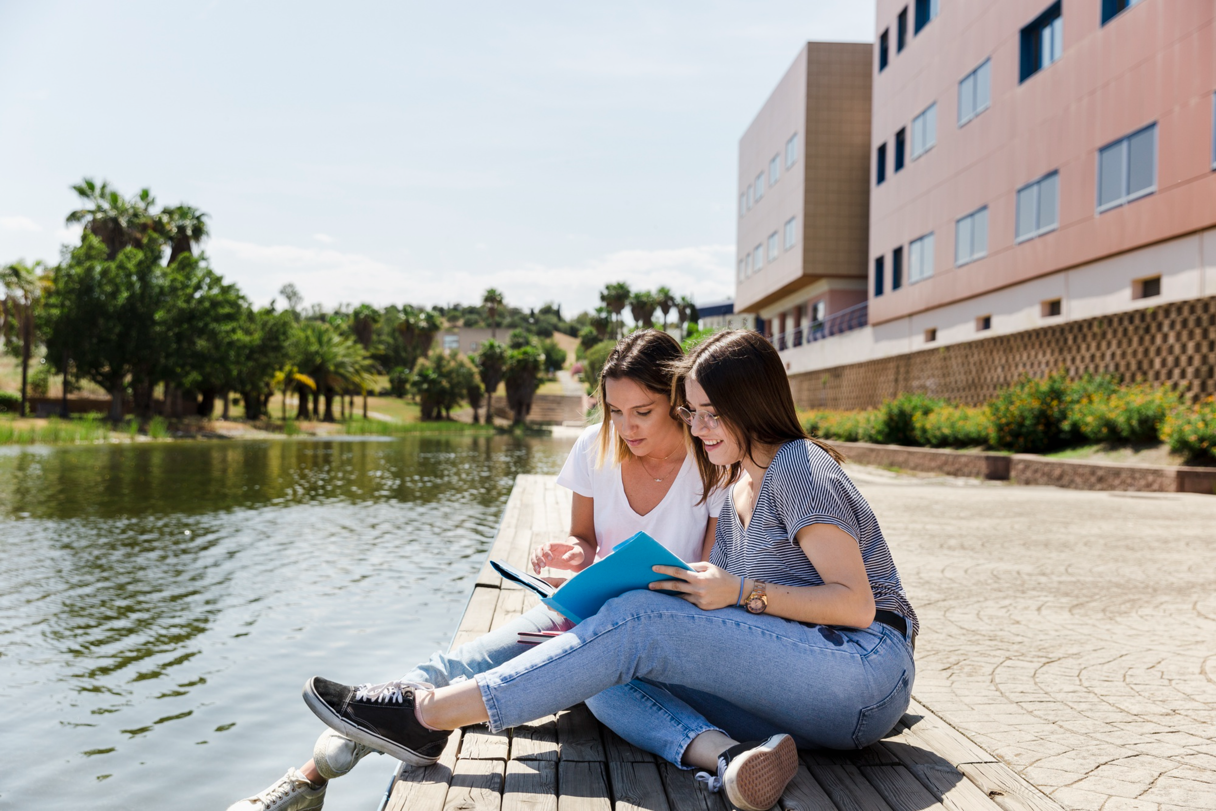 Two young women sitting on a wooden dock by a lake, looking at a book together, with a modern building and trees in the background on a sunny day.