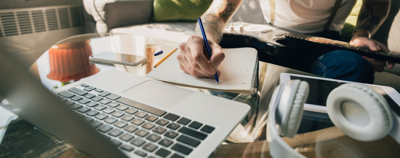 Person writing in notebook with laptop, tablet, headphones, and smartphone on glass table