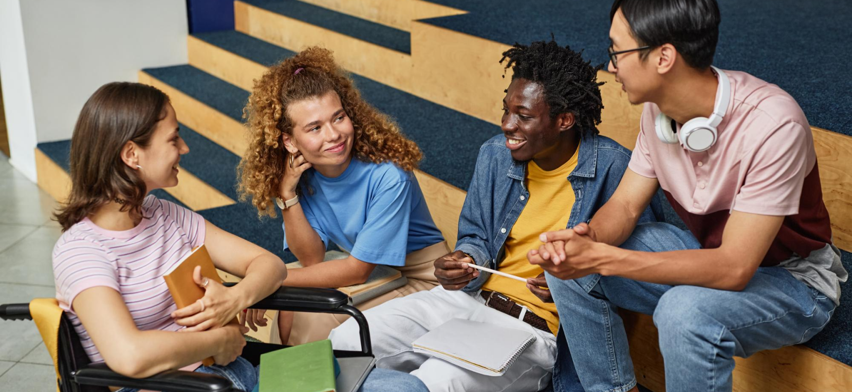 Group of four young people sitting on stairs, engaging in conversation, with notebooks and a smartphone, in a casual setting.