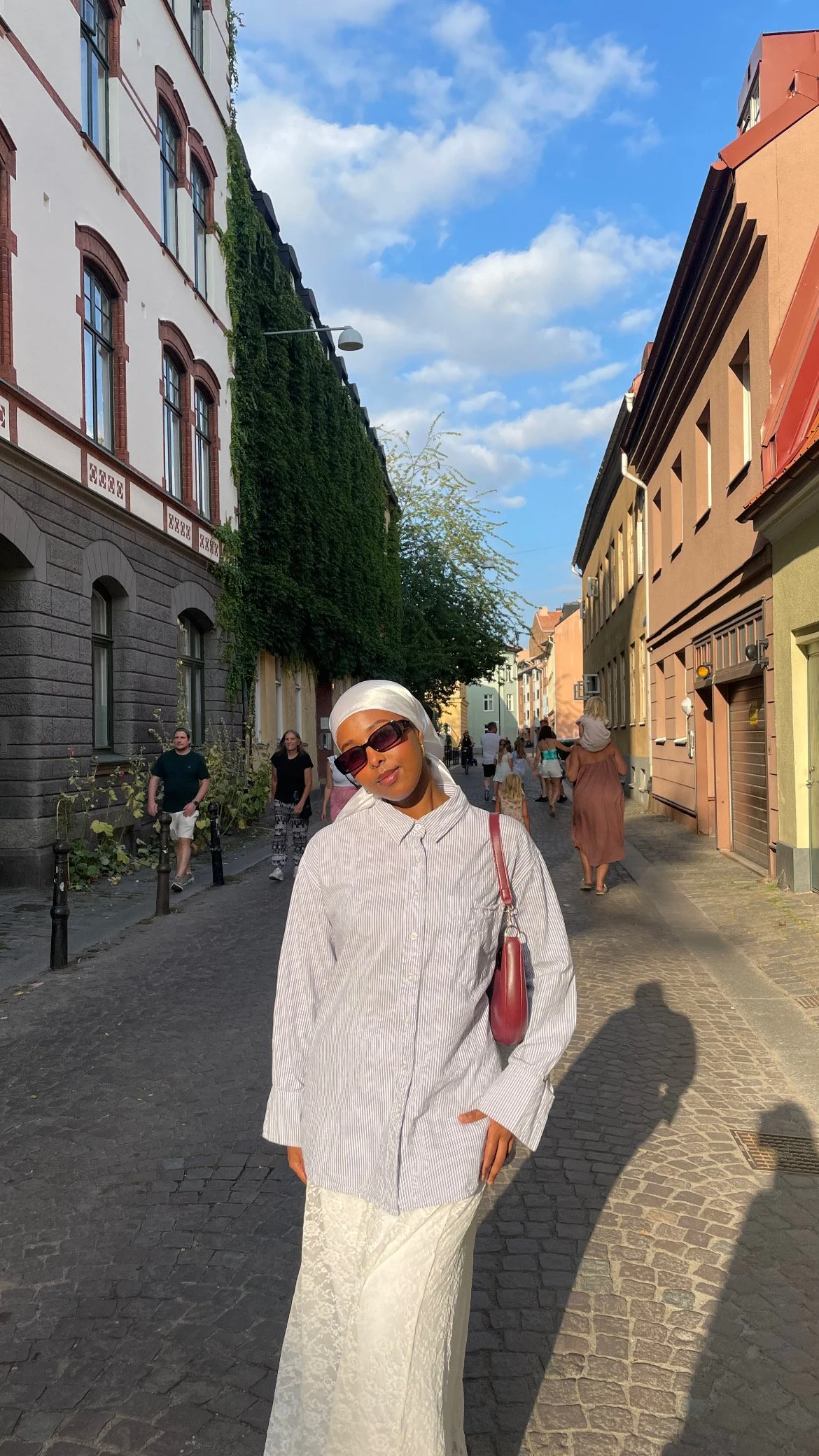 A woman in a striped shirt, white skirt, and sunglasses standing on a cobblestone street in a European city, with colorful buildings and other pedestrians in the background.
