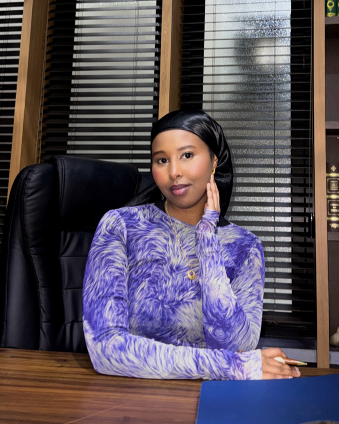 A woman sitting at a wooden desk in an office, wearing a purple and white patterned top, with a black headscarf, in front of window blinds.