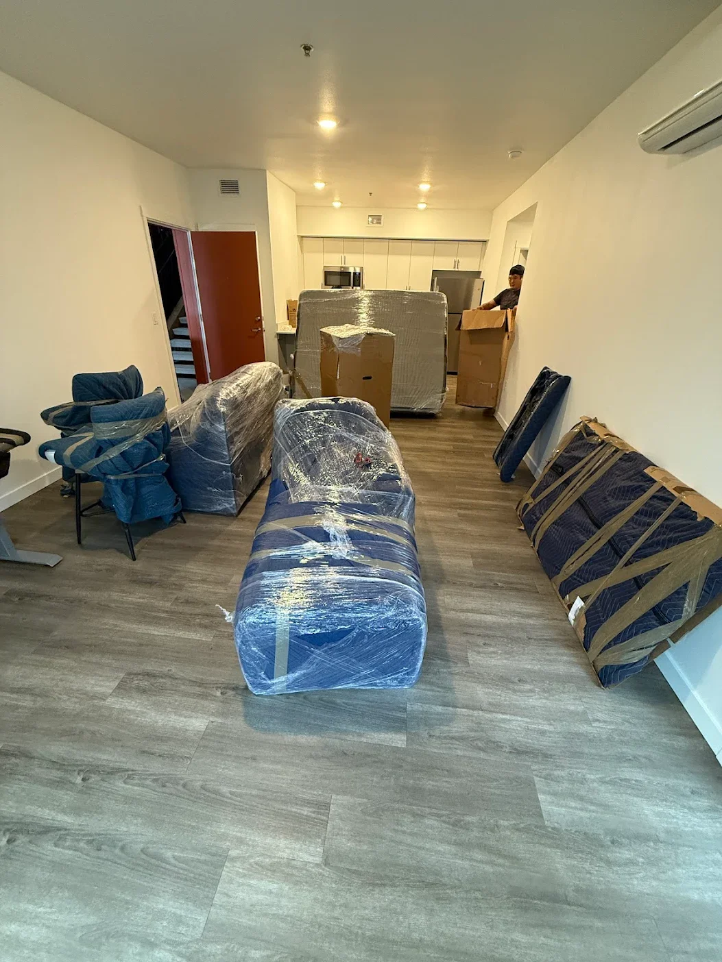 Living room with furniture wrapped in plastic, moving boxes, and packaging tape, indicating moving or unpacking activity.