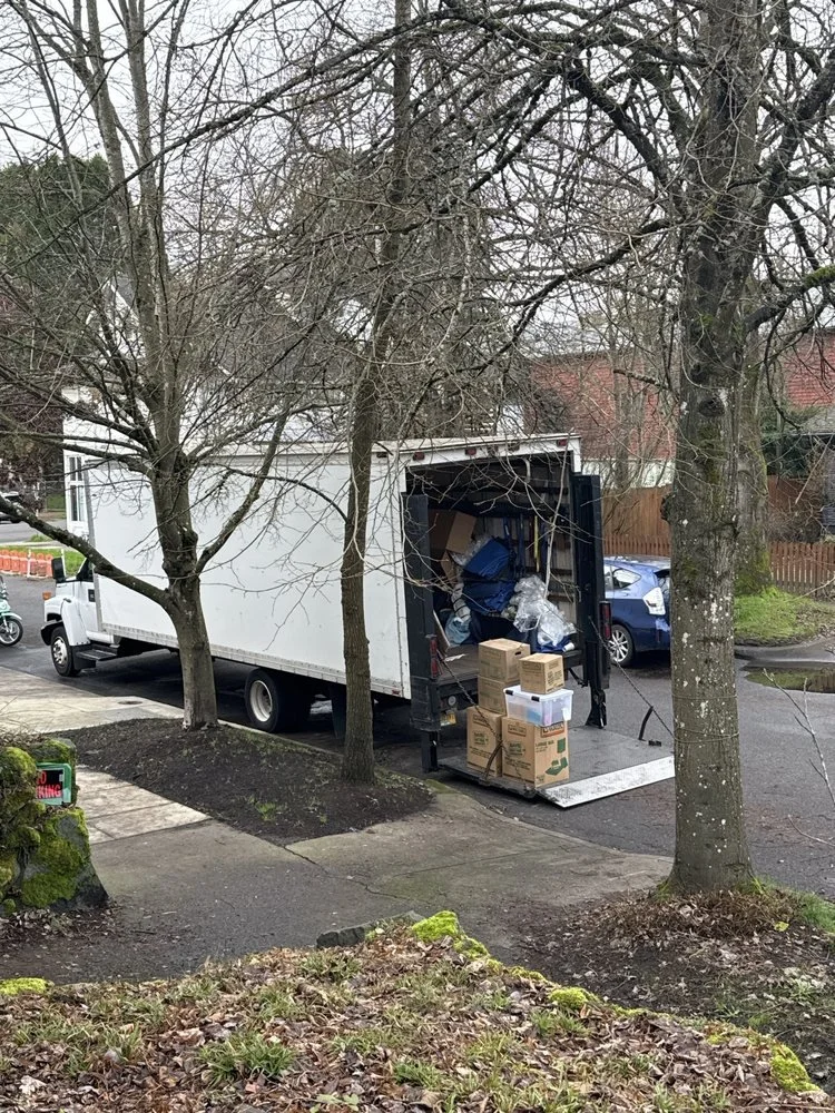 A moving truck parked on the street with its back open, filled with boxes and other moving supplies. Several boxes are placed on the ground at the truck's entrance. The scene is in a neighborhood with leafless trees and nearby parked cars.