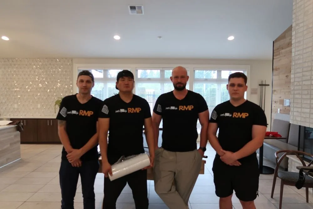 Four men standing indoors, wearing matching black RMP t-shirts, posing for a photo in a bright room with large windows and modern decor.