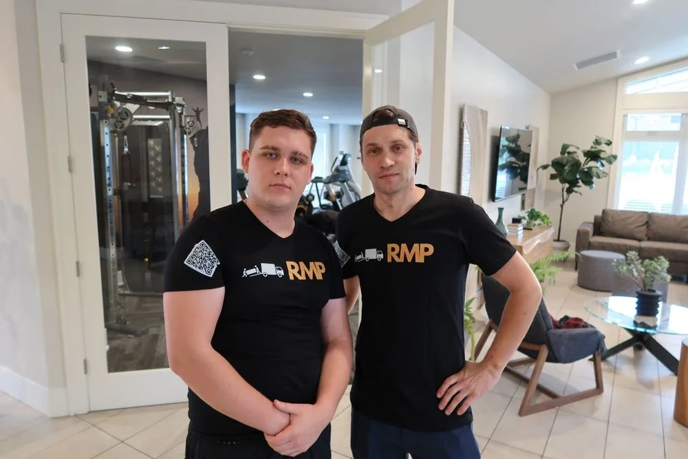 Two young men wearing black t-shirts with a tow truck graphic and 'RMP' logo, standing in a bright living room with modern furniture and a TV mounted on the wall.