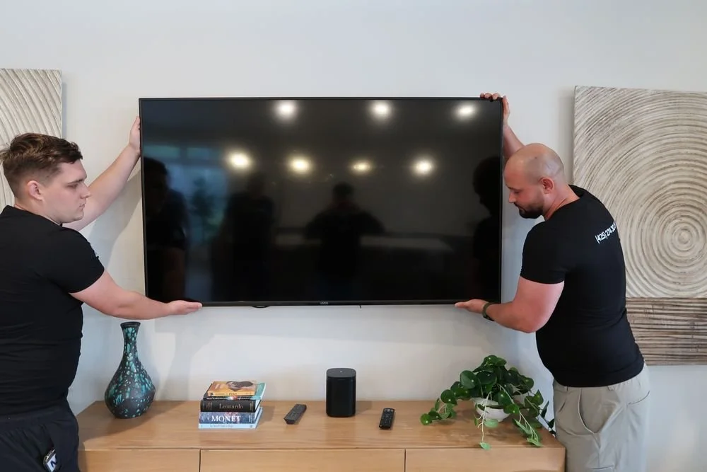 Two men are mounting a flat-screen TV on a wall in a living room. The TV is centered above a wooden console with decorative items, including a vase, books, a speaker, remote controls, and a potted plant.