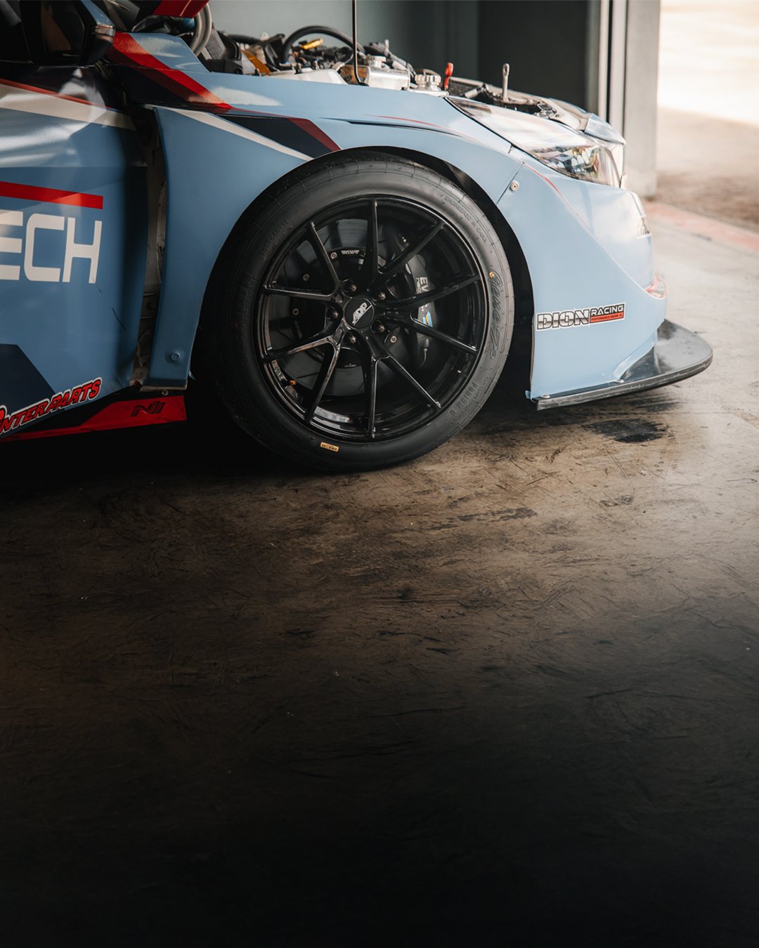 Close-up of a race car's front section with black alloy wheels, blue bodywork, and racing decals, parked on a concrete floor near a garage.