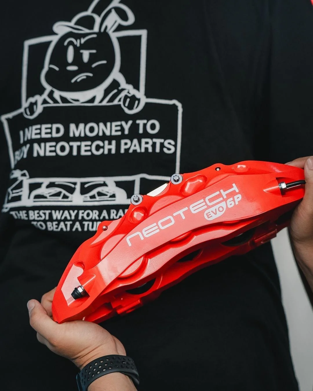 Person holding a red NeoTech Evo 6 Piston brake caliper in front of a person wearing a black shirt with white graphic text about Neotech parts.