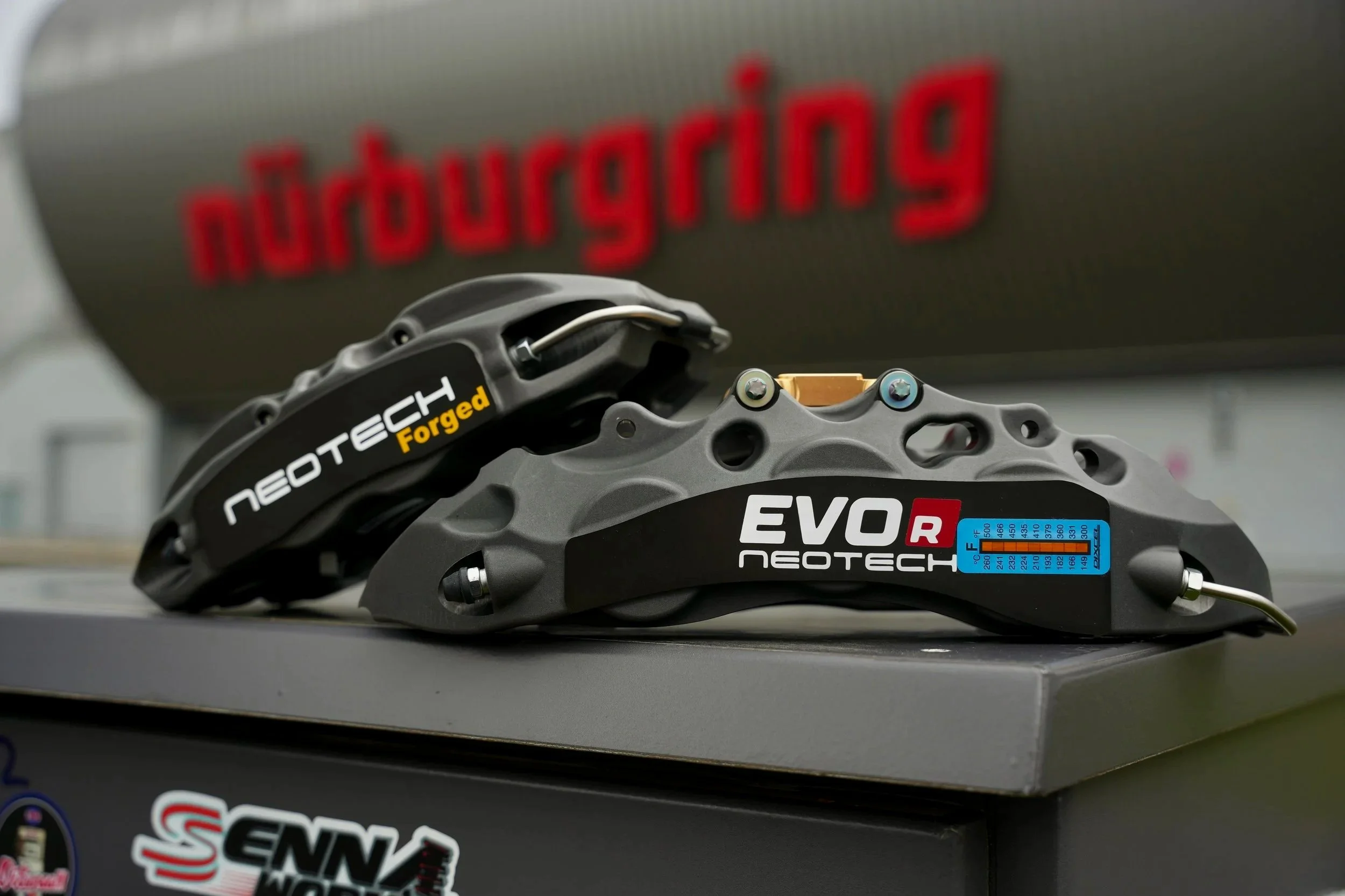 Close-up view of black Neotech EVO R brake calipers with branding, placed on a gray surface, with a blurred background showing an Nurburgring sign.