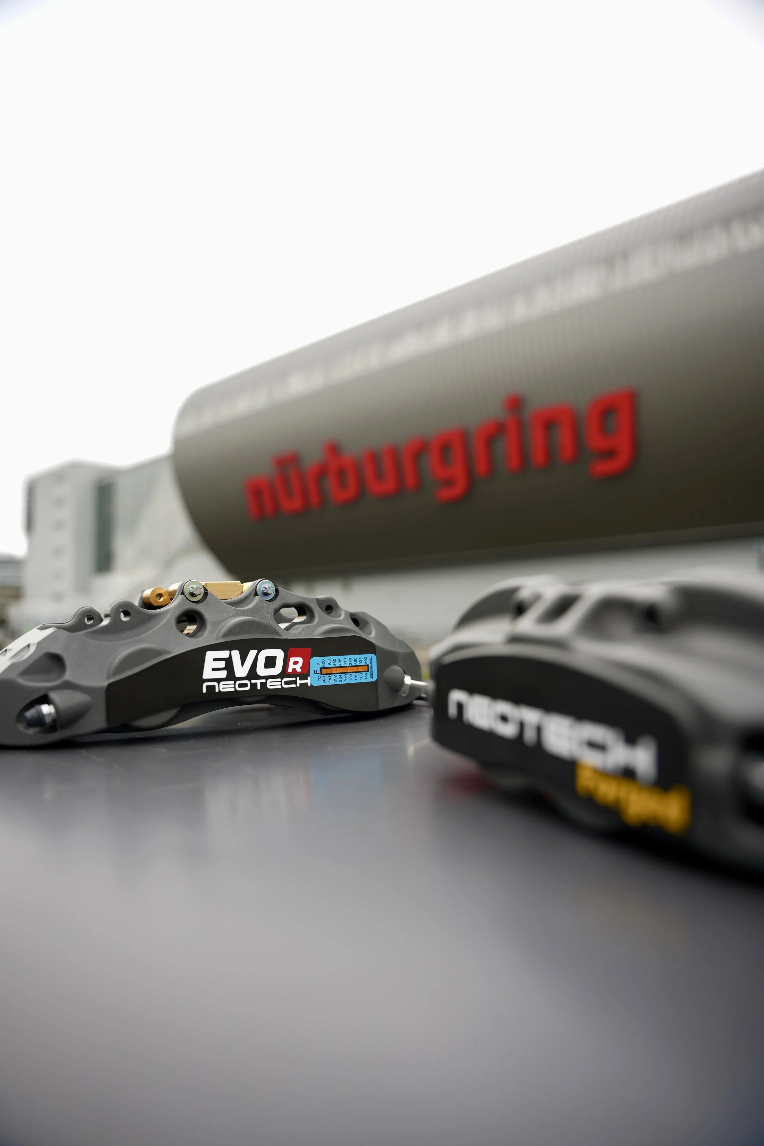 Close-up of two motorcycle brake calipers, one labeled 'EV2 R' and 'NEOTECH', on a surface in front of a building with a sign that reads 'nürburgring'.