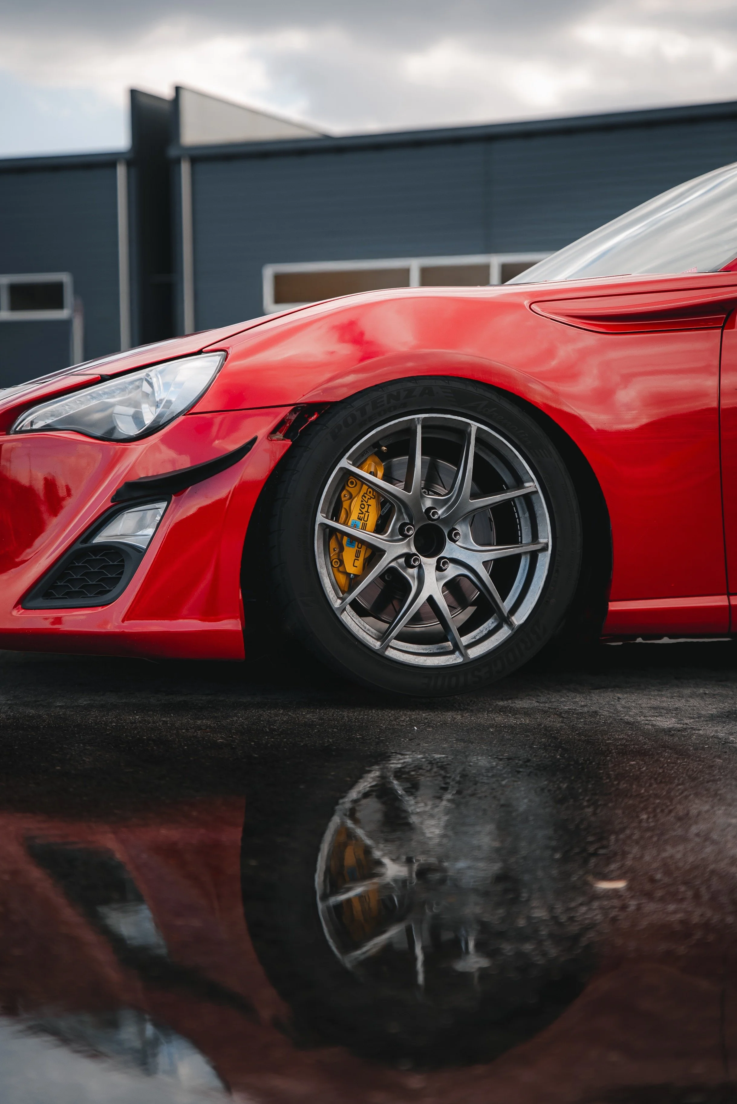 Close-up of a red sports car with a visible yellow brake caliper, parked on wet ground with its reflection visible, and a modern building in the background.