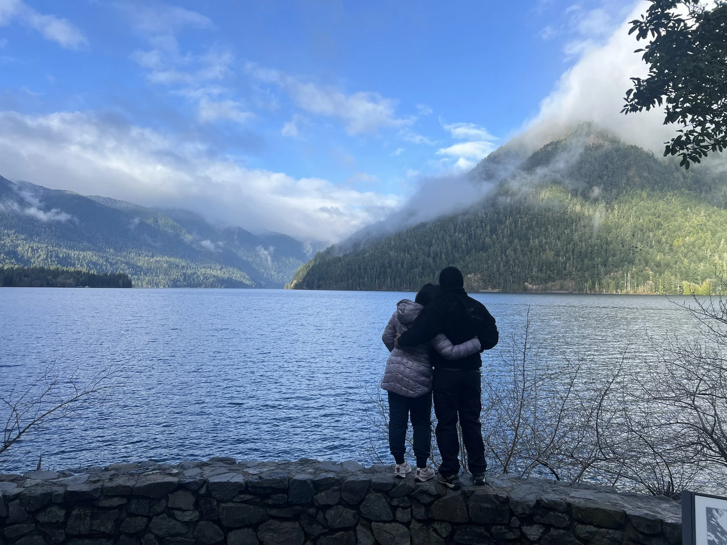 Two people wearing jackets with arms around each other, standing on a stone wall by a lake with forested mountains in the background, under partly cloudy skies.