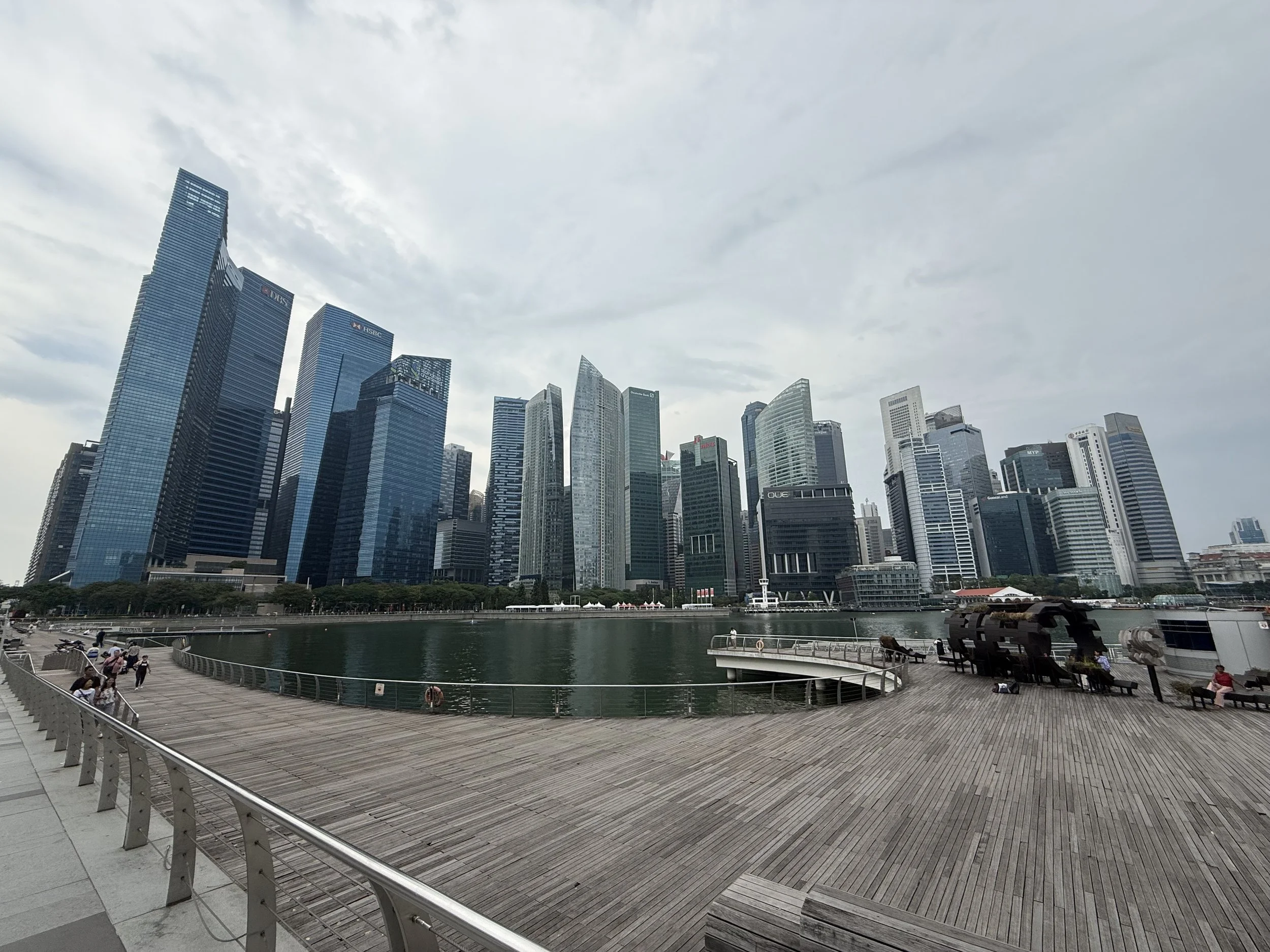 City skyline with tall modern skyscrapers behind a water body and a wooden deck with benches and people sitting or walking.