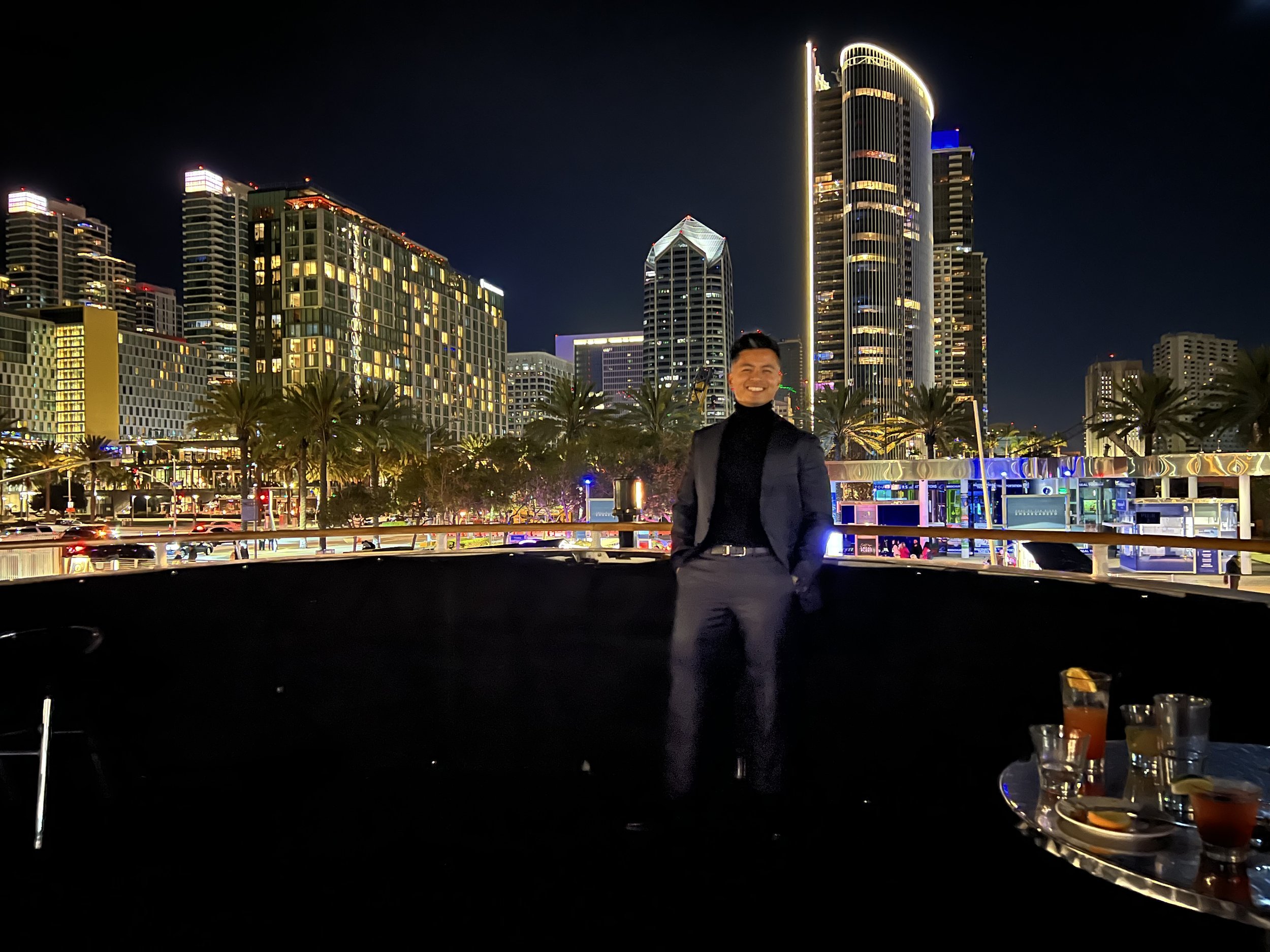 A man in a black suit and turtleneck standing on a boat at night, with a city skyline illuminated by lights in the background.