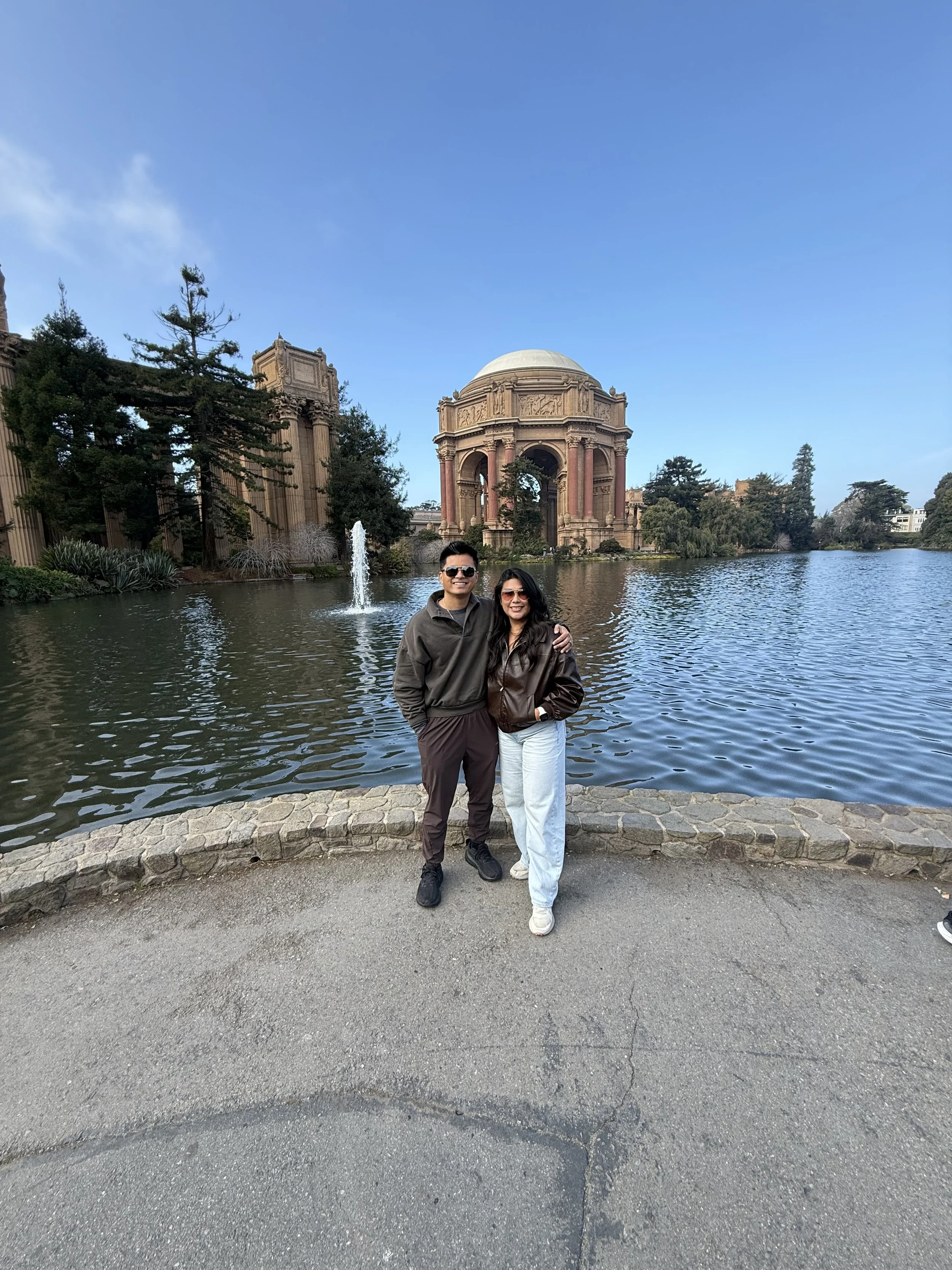 A man and woman standing together by a lake with the Palace of Fine Arts in San Francisco in the background, smiling and posing for a photo on a clear day.