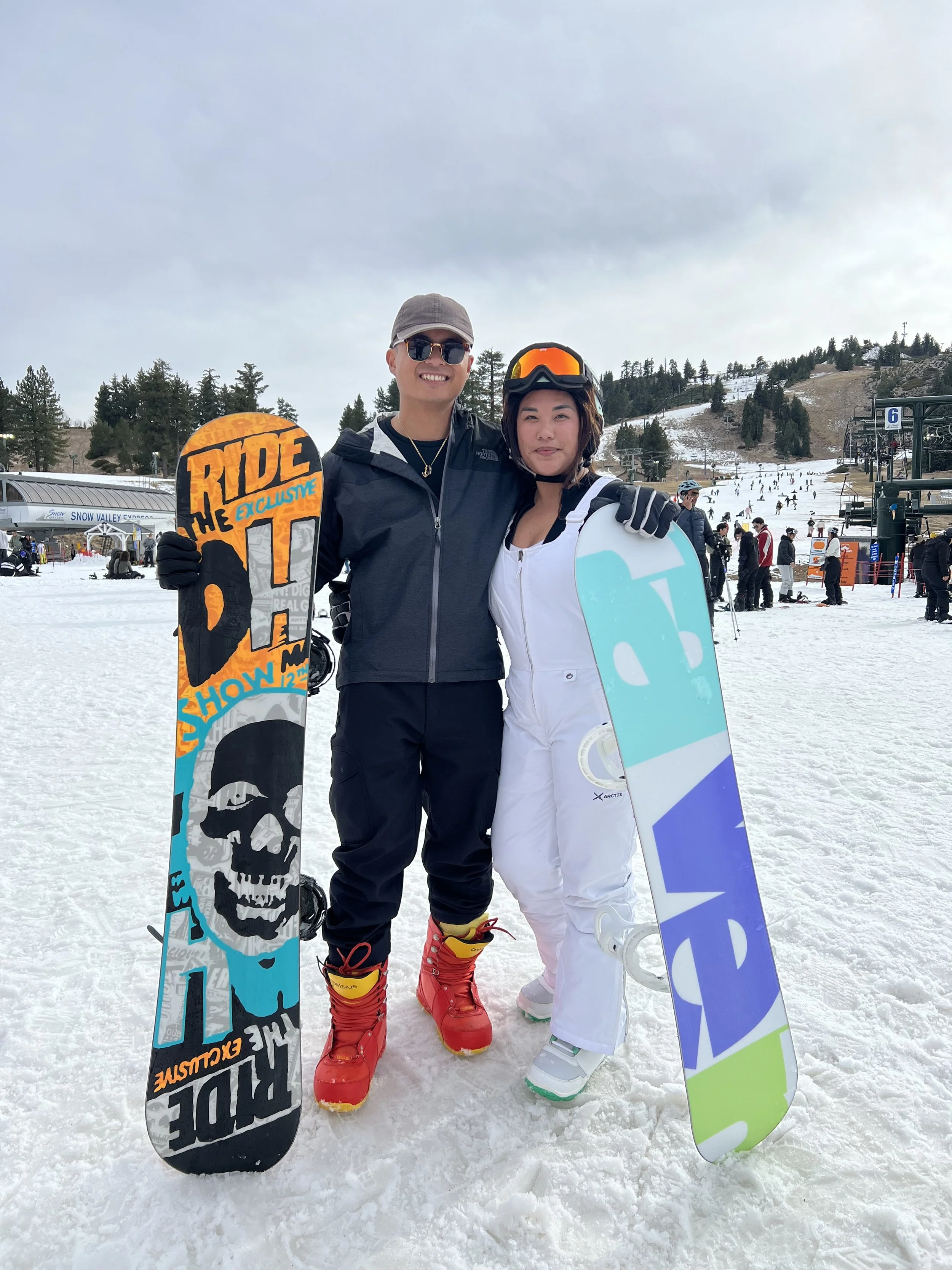 Two people in ski gear standing on snow at a ski resort, holding snowboards, with ski lifts and other skiers in the background.