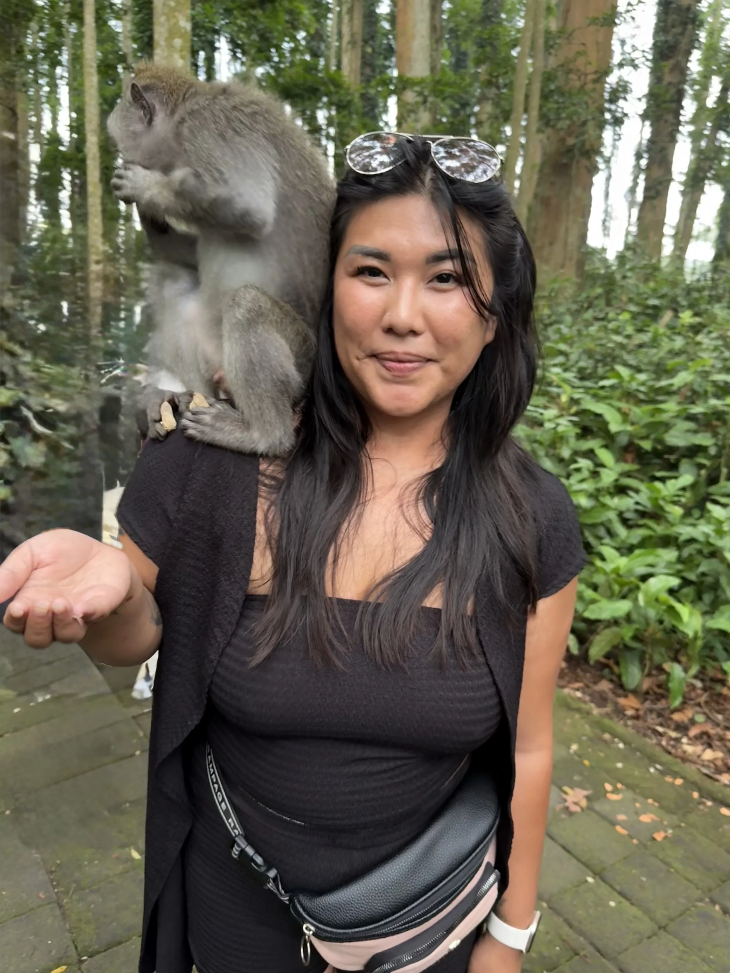 A woman with sunglasses on her head standing outdoors with a monkey on her shoulder in a forested area.