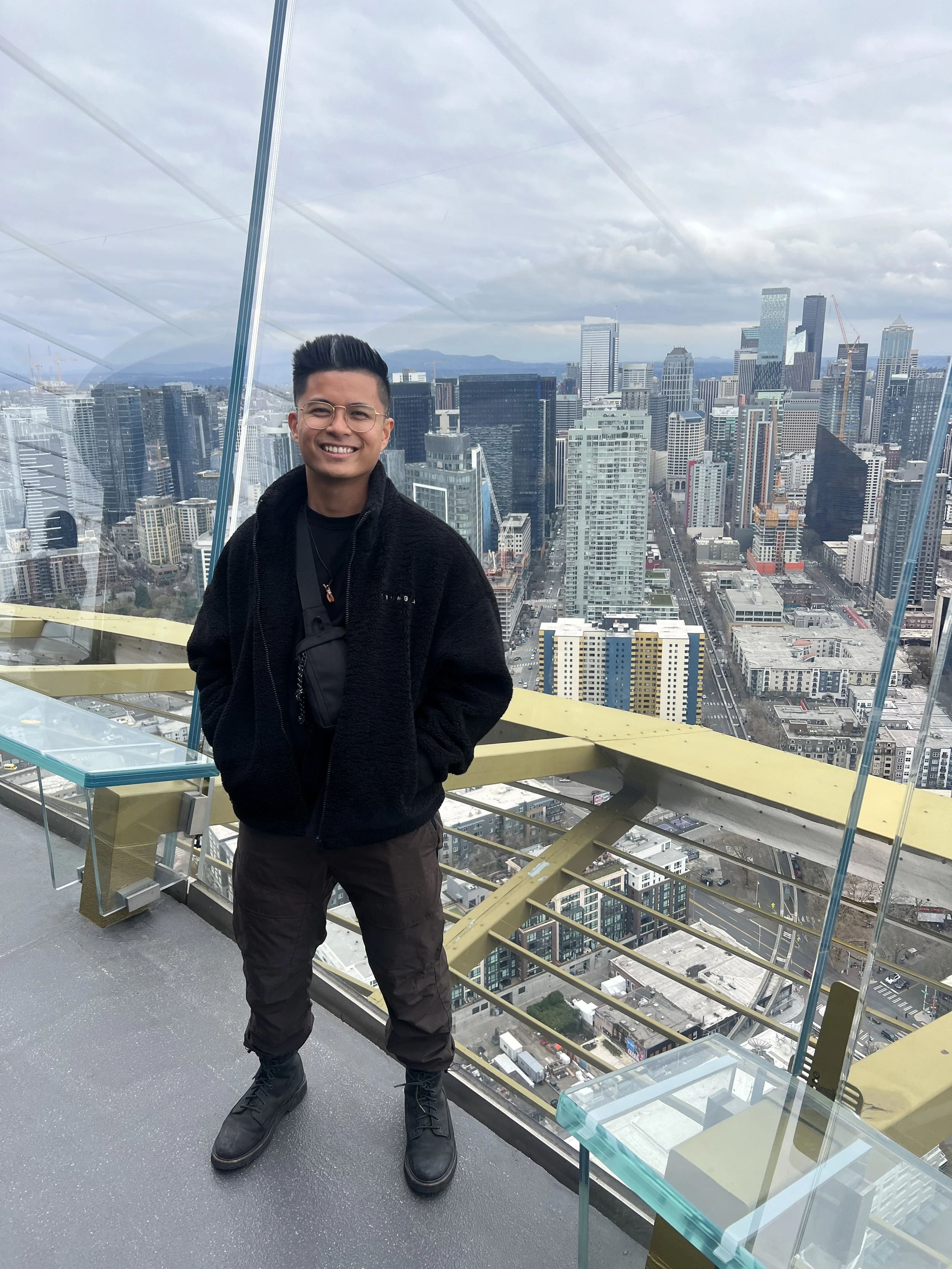 Young man smiling on a rooftop viewing platform with a city skyline and tall buildings in the background.