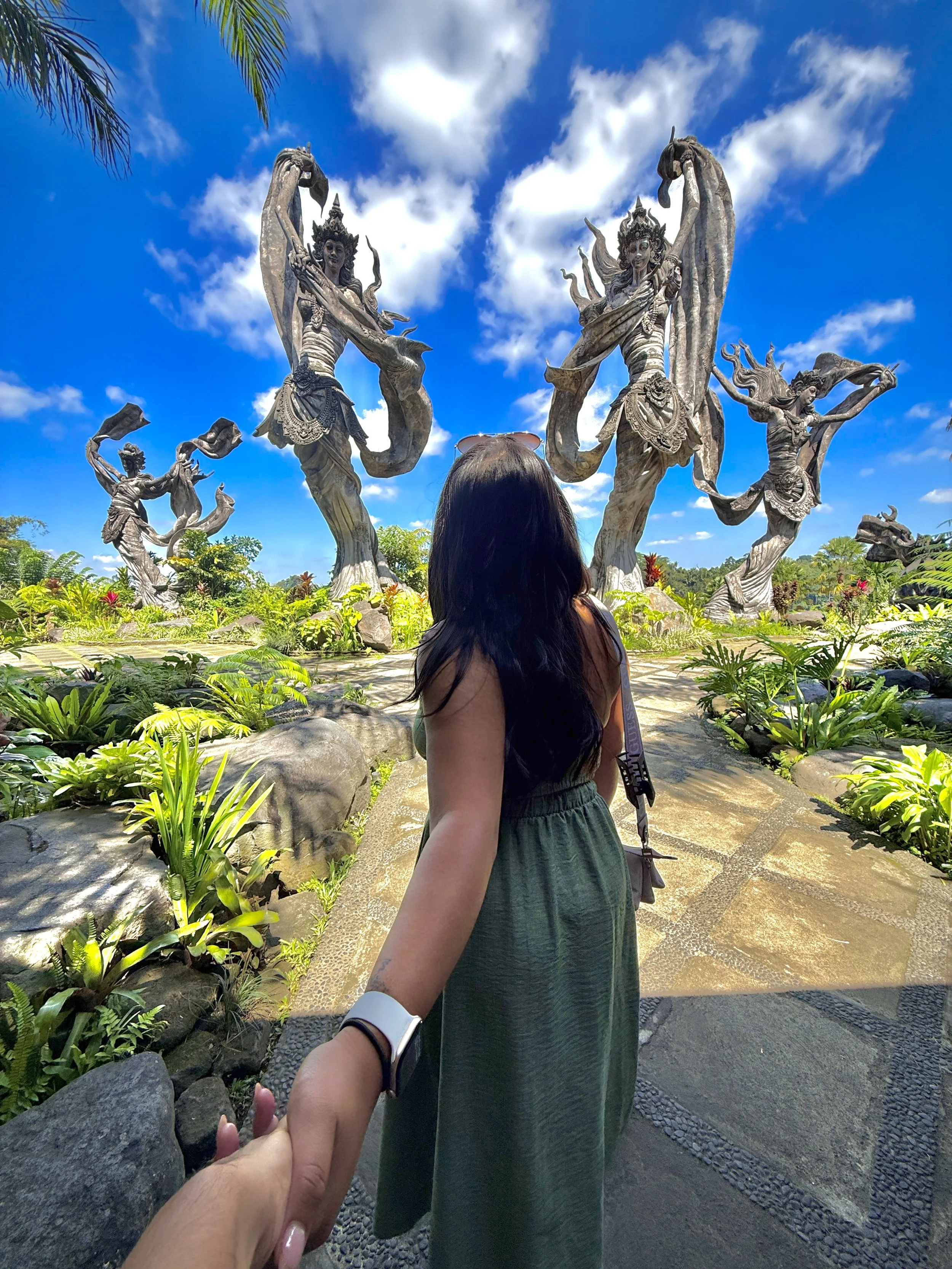 A woman holding another person's hand at a tropical outdoor location with large sculptures of mythological figures in the background, under a partly cloudy blue sky.