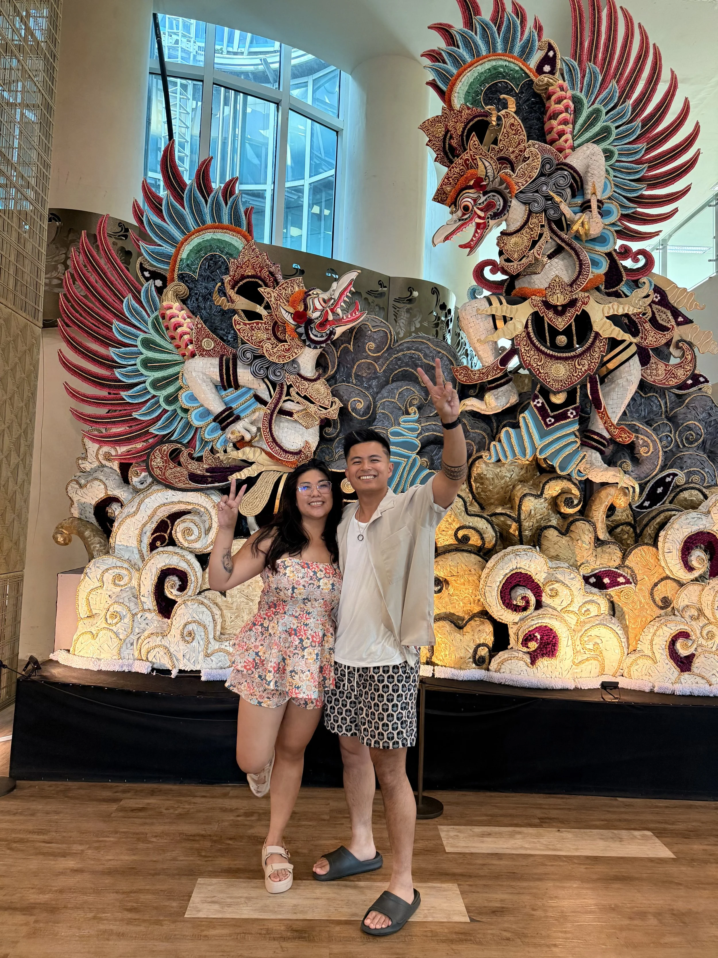 Two young adults, a woman and a man, pose happily in front of a large colorful ornate sculpture of mythical creatures, likely dragons or phoenixes, decorated with vibrant fabrics and intricate patterns, inside a modern building with glass walls.