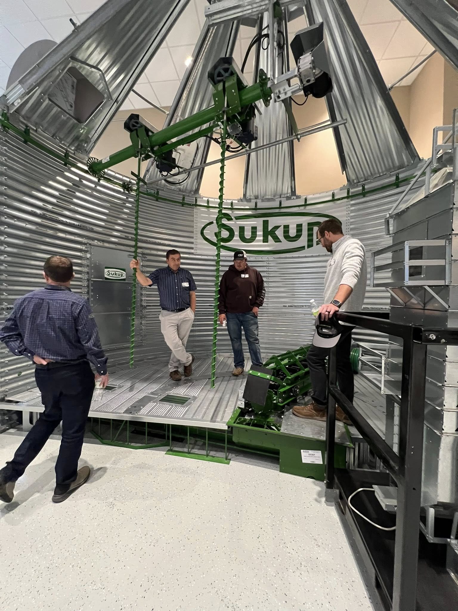 Group of four men inside a large metallic silo with the word 'SUKUP' on the wall, examining equipment including two large green mechanisms and a vertical spiral auger.