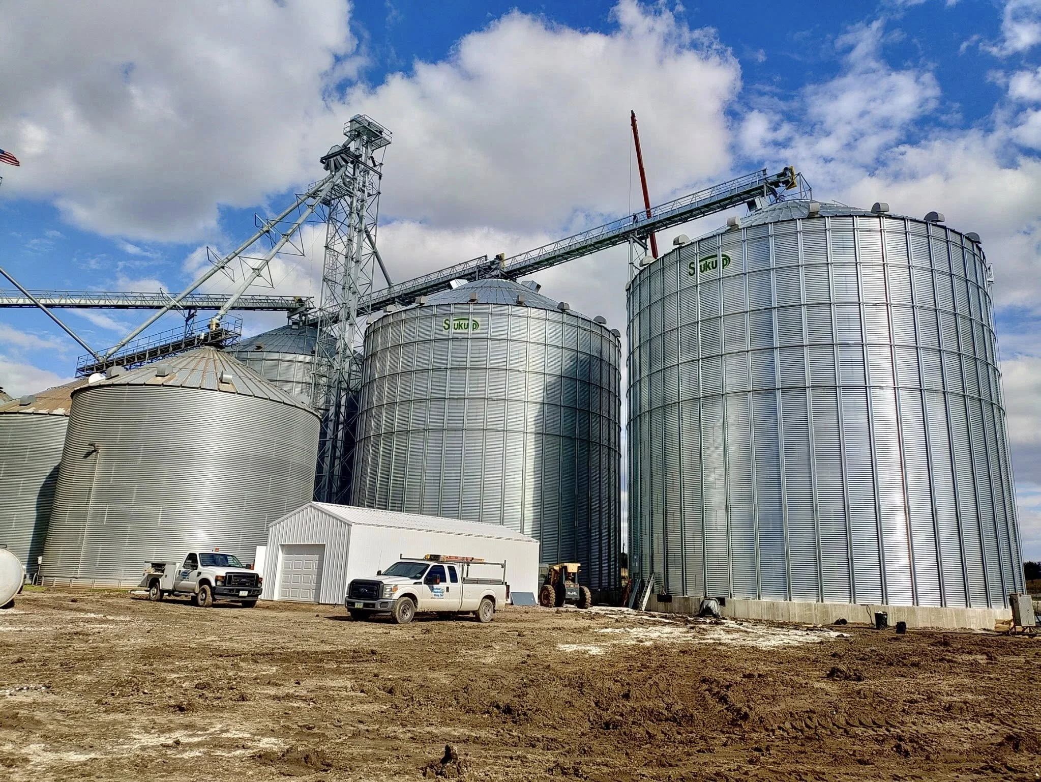 Large metallic grain silos with a crane on top under a partly cloudy sky, with trucks and a small building in front on barren ground.