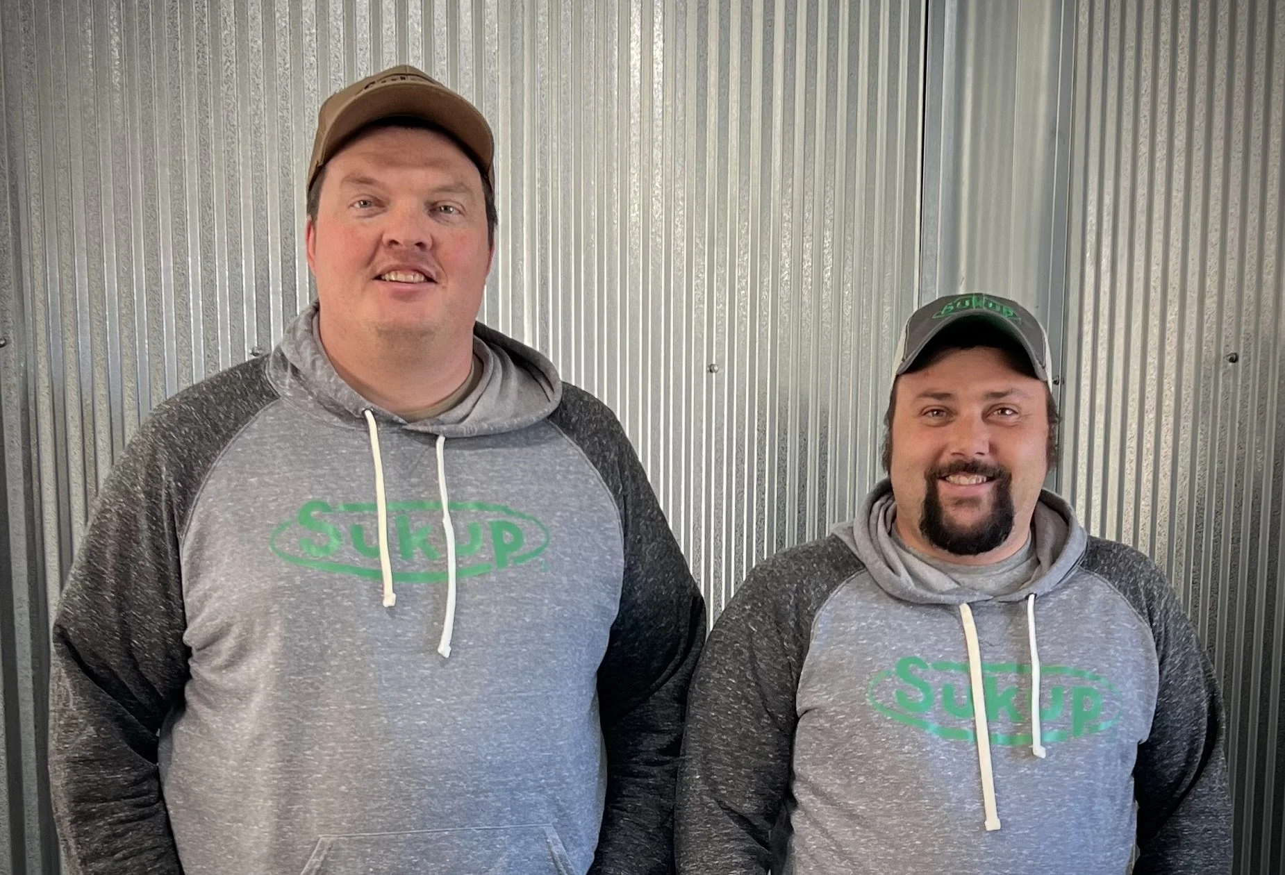 Two men standing in front of a metal wall, both wearing gray and black hooded sweatshirts with the word 'SUKUP' in green on the front, and gray caps. They are smiling at the camera.