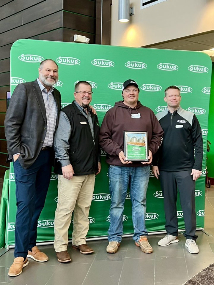 Four men standing in front of a green backdrop with the Sukup logo, one holding a framed award or certificate, inside a modern building with dark wood paneling and lighting.